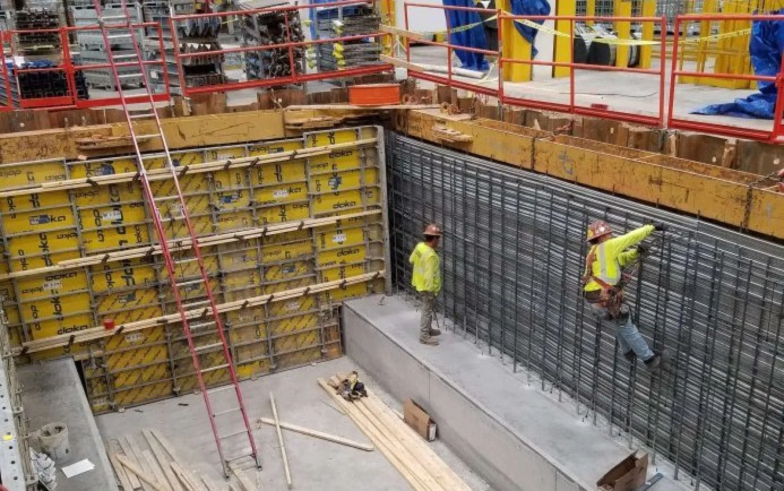 Construction workers in safety gear installing steel reinforcement inside a concrete foundation with yellow formwork panels.