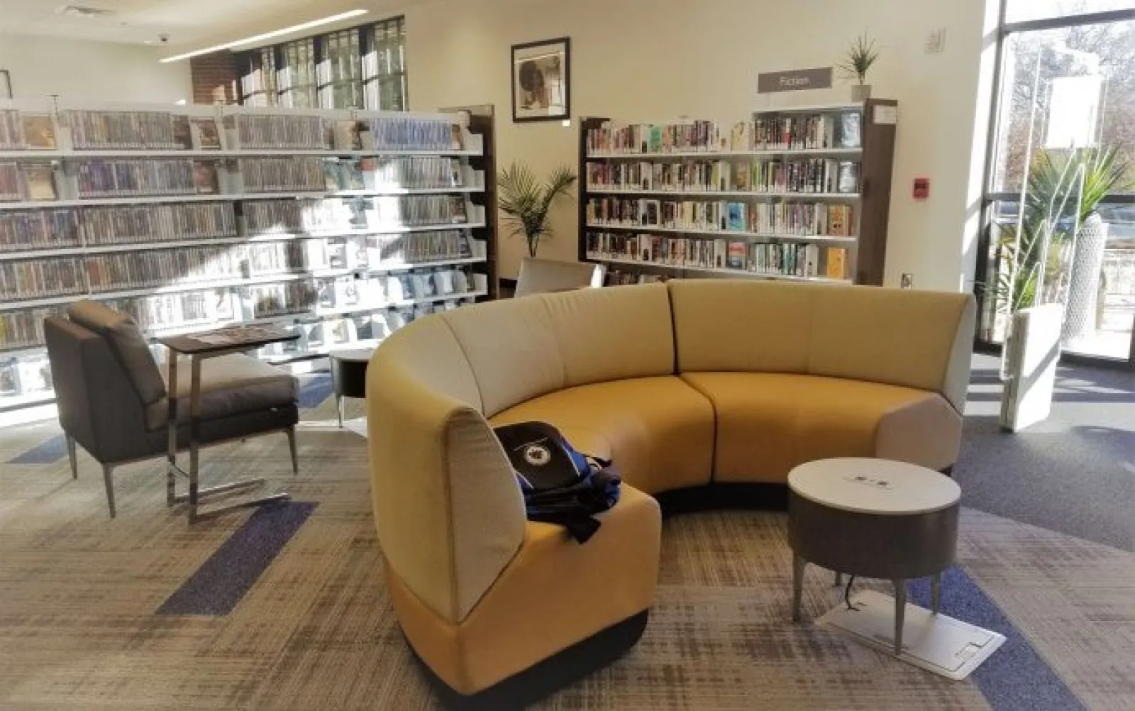 Modern library interior with curved beige sofa, bookshelves, armchair, and large windows letting in natural light