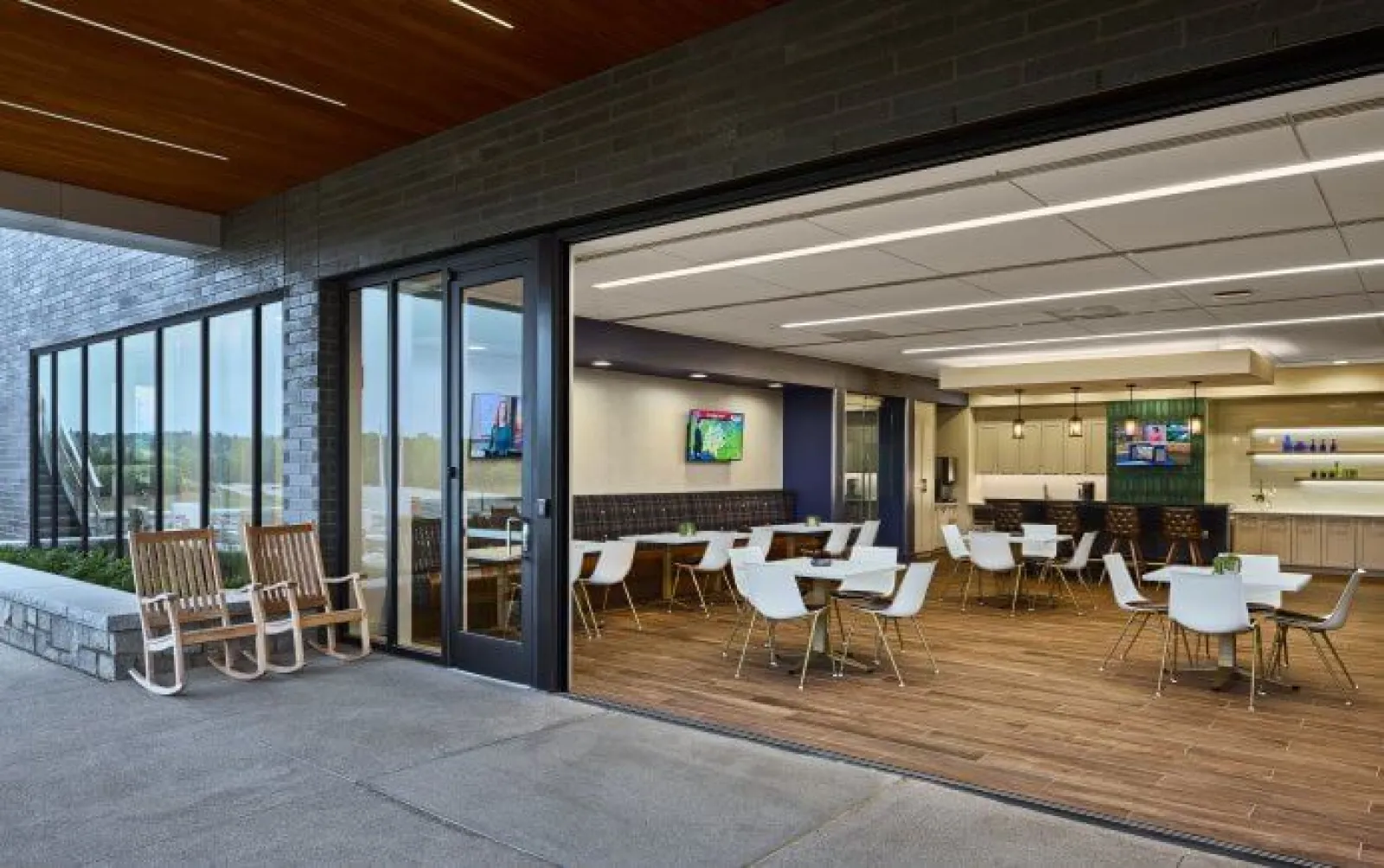 Modern indoor dining area with white chairs and wood flooring opens to an outdoor patio with rocking chairs.