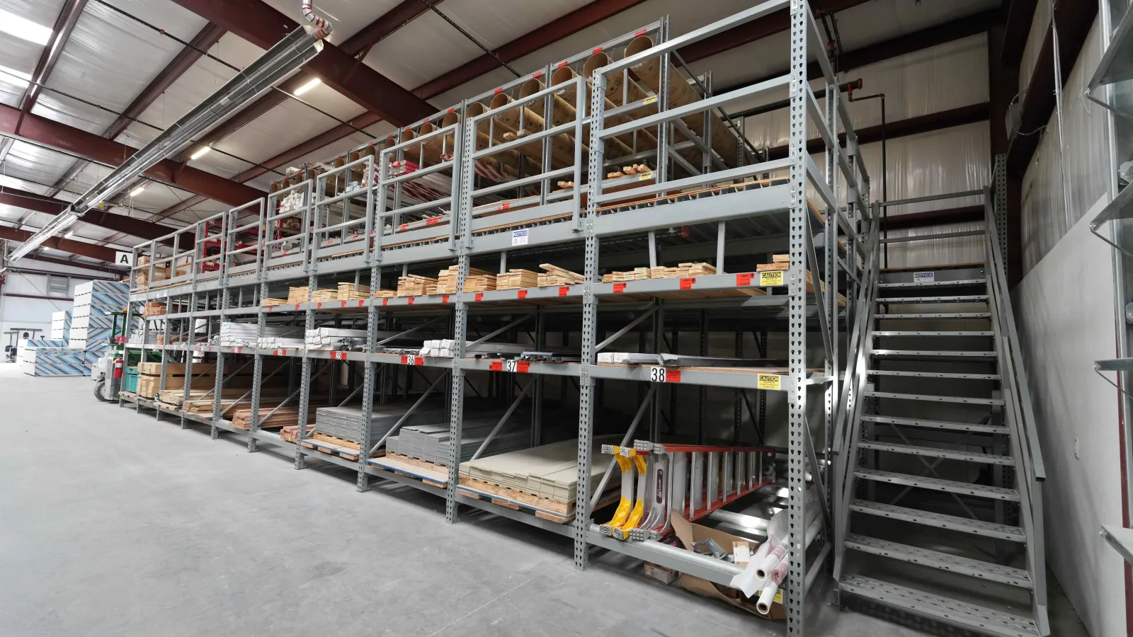 Industrial warehouse metal shelving with wooden pallets, construction materials, and a staircase alongside the racks.