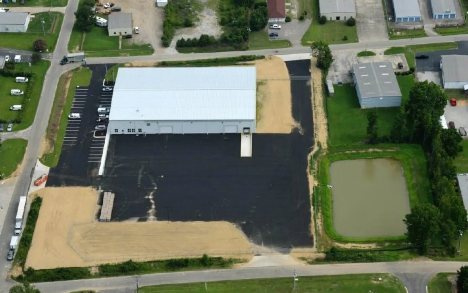 Aerial view of industrial warehouse with large parking lot and adjacent pond surrounded by greenery and roads.