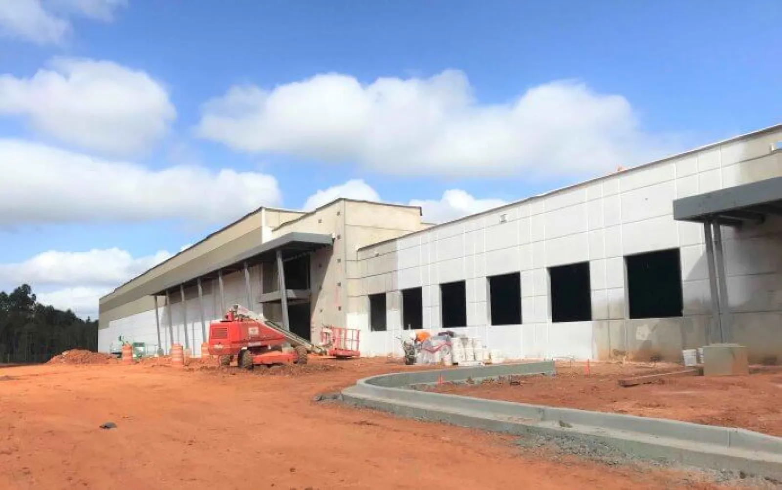 Construction site with a large industrial building under development and orange dirt ground under a blue sky.