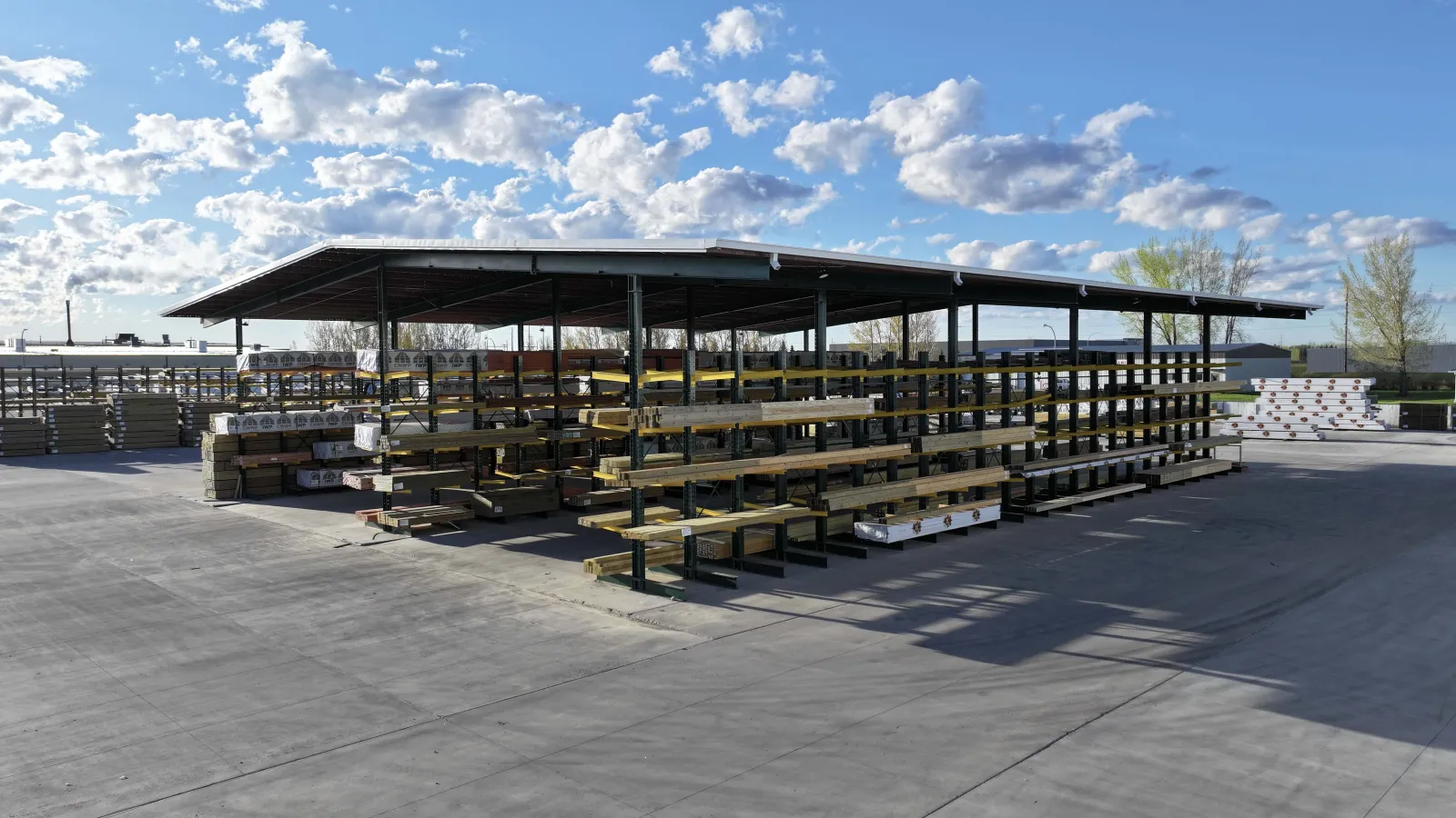 Outdoor lumber storage with organized metal racks under a metal roof on a sunny day with scattered clouds.