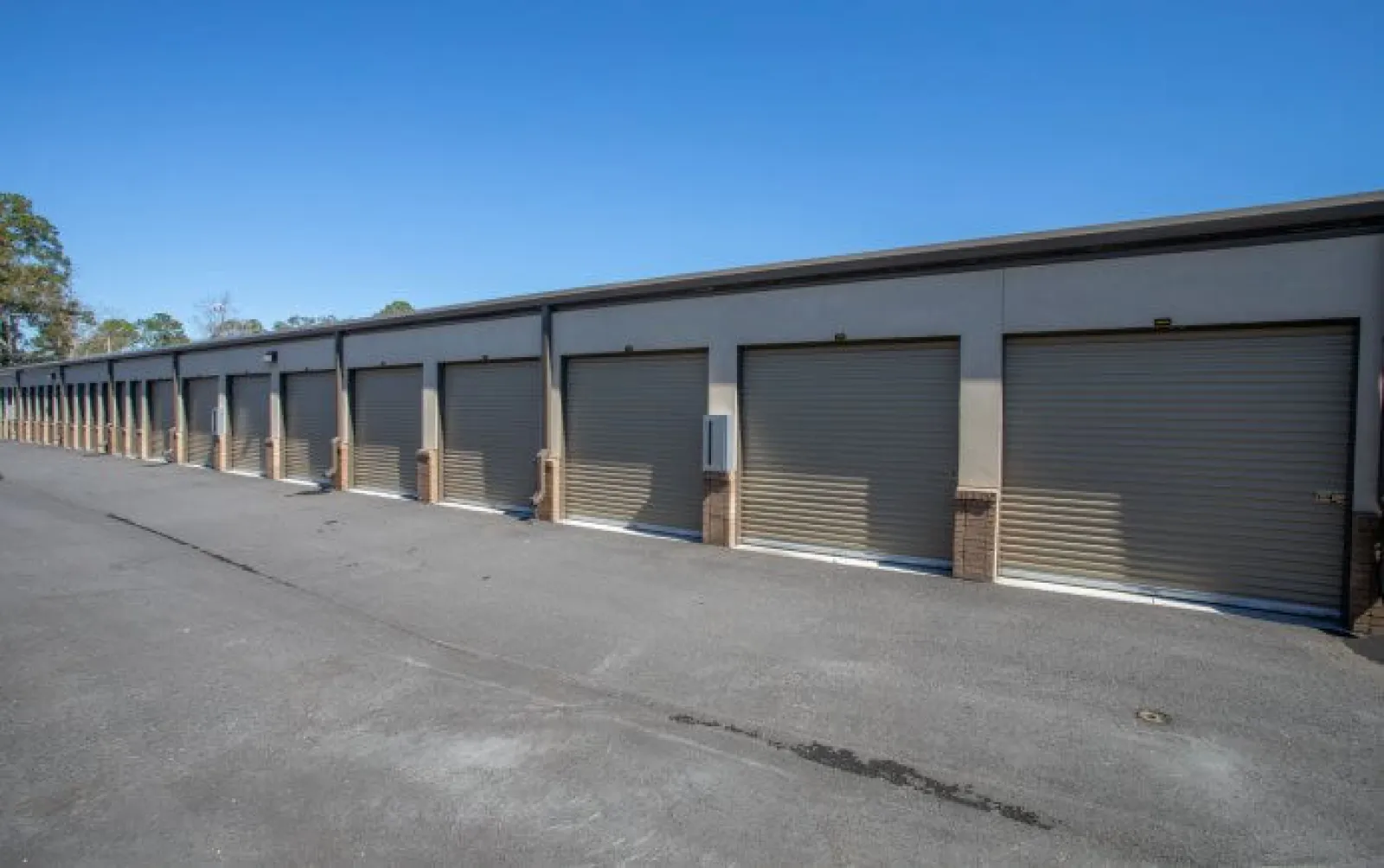 Row of closed beige storage unit doors along a paved driveway under clear blue sky.