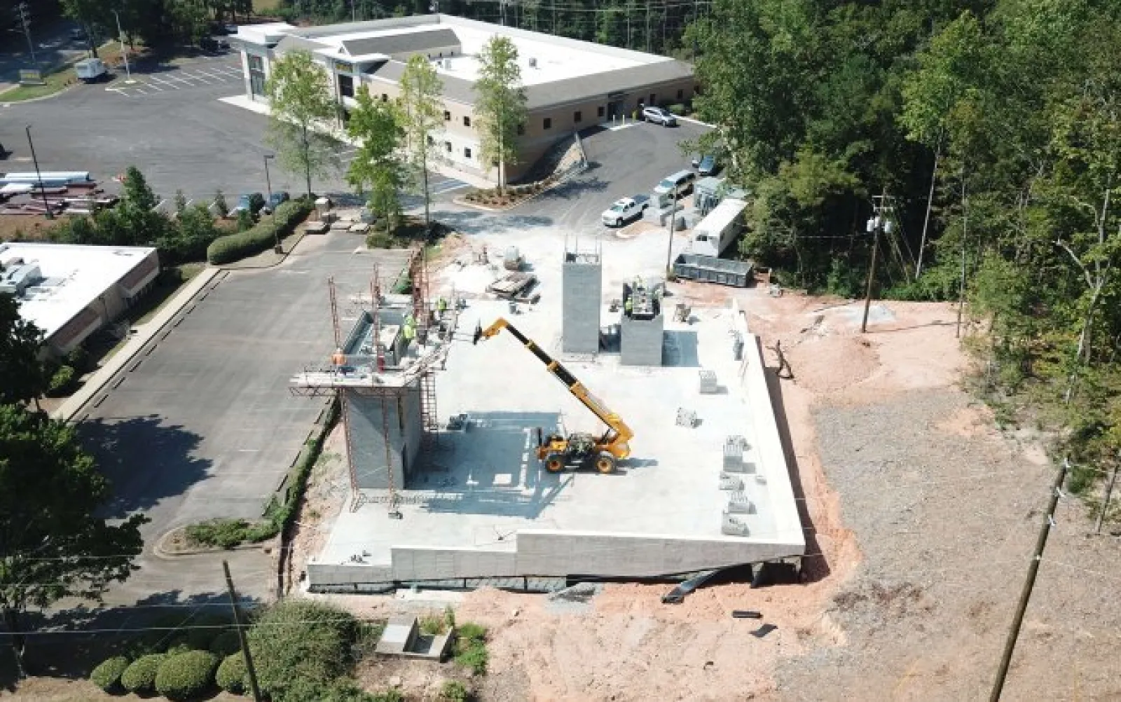 Aerial view of a construction site with concrete foundation, crane, and workers amidst surrounding trees and buildings.
