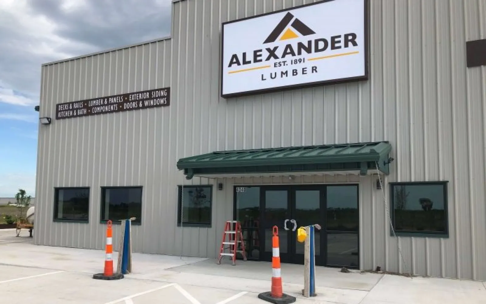 Front view of Alexander Lumber building with signage, traffic cones, and large windows on a cloudy day