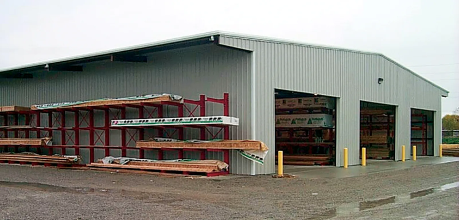 Exterior view of a metal storage building with open bays and lumber racks on a gravel lot