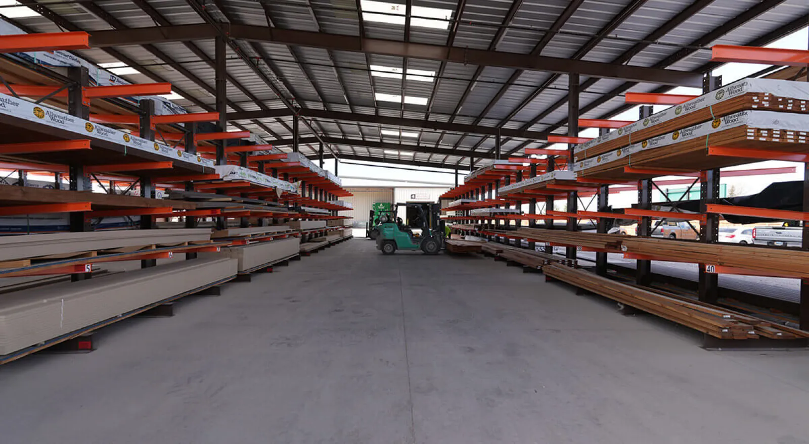 Warehouse interior with metal and wood materials stored on orange racks and forklifts in the center aisle.