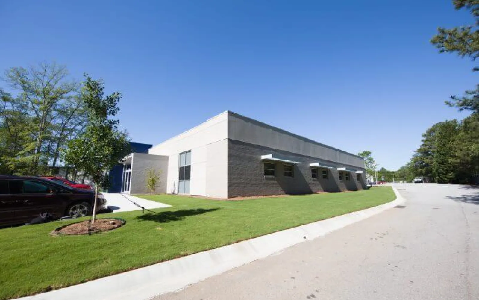 Modern single-story commercial building with gray walls, green lawn, parked cars, and clear blue sky.