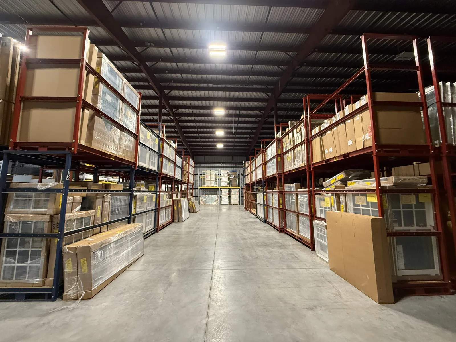 Spacious warehouse aisle with tall red metal racks filled with cardboard boxes and packaged goods under bright ceiling lights