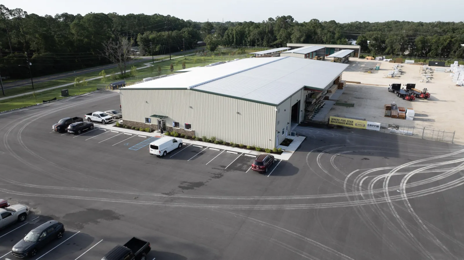 A large industrial warehouse with several parked vehicles and tire marks in a spacious asphalt lot surrounded by trees.