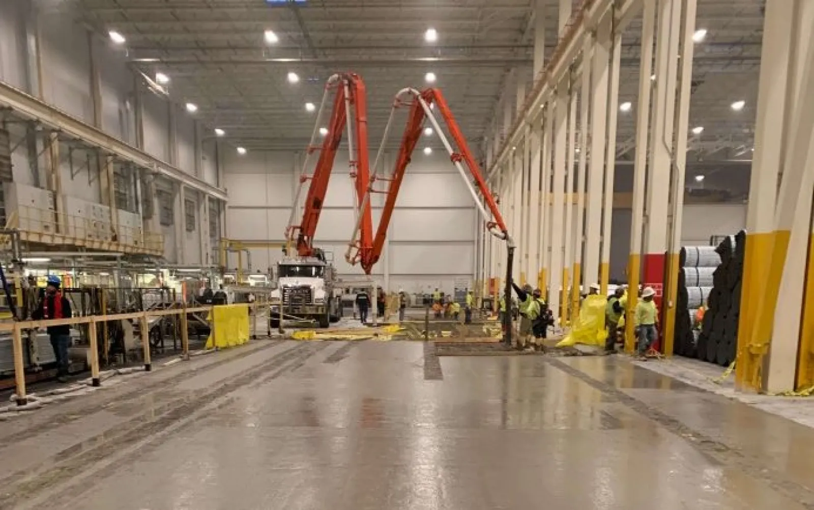 Workers using concrete pump trucks inside a large industrial warehouse with high ceilings and pillars.