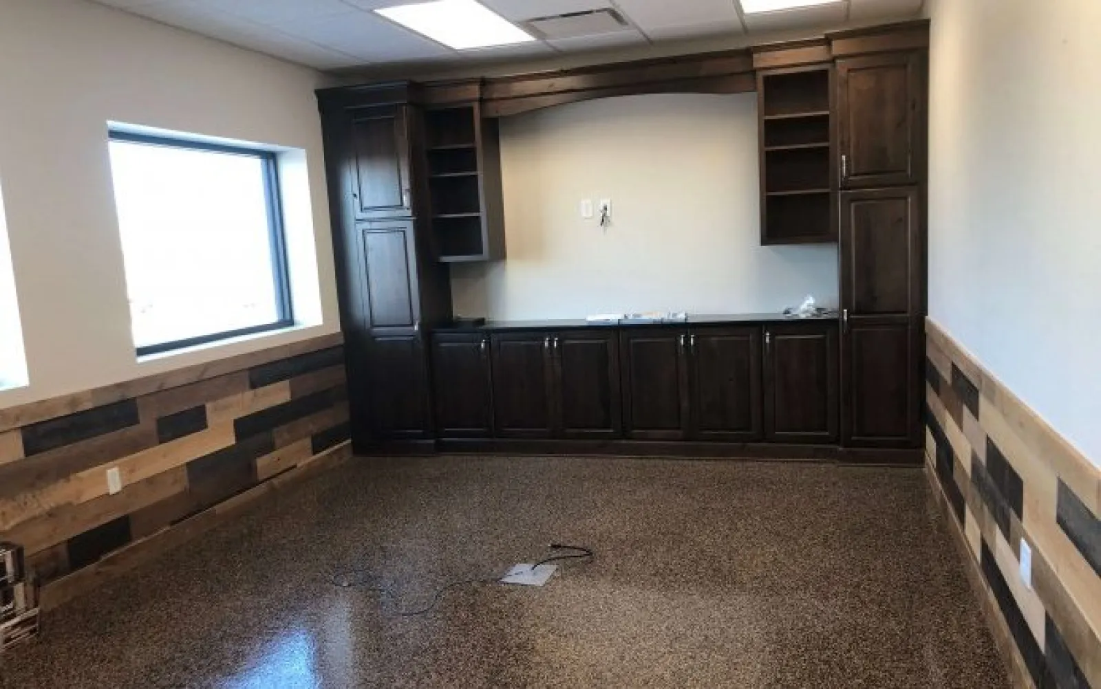 Empty room with polished concrete floor, wood panel walls, large window, and dark wooden cabinets against the wall.
