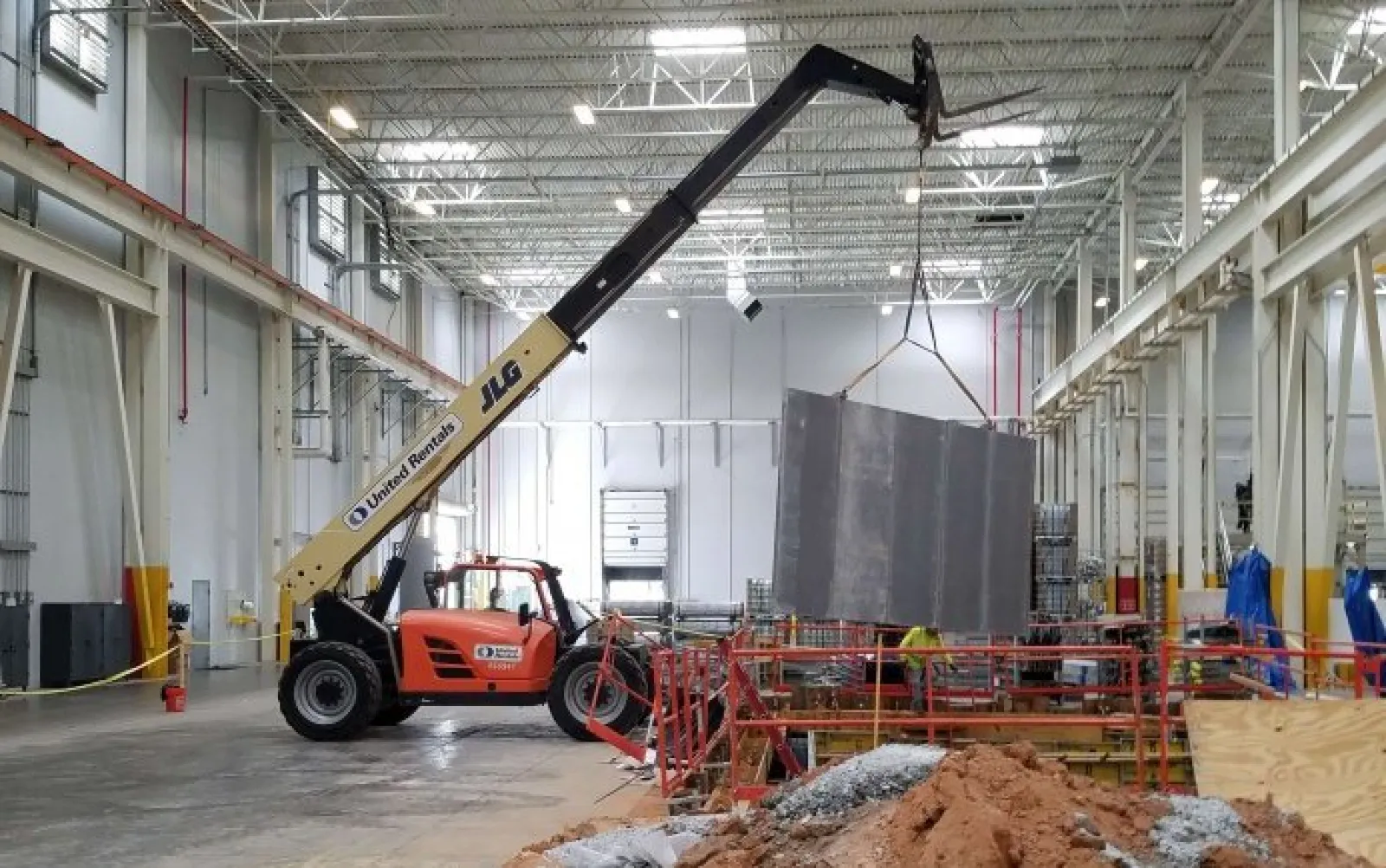 Orange JLG telehandler lifting a large metal panel inside a spacious industrial warehouse under construction.