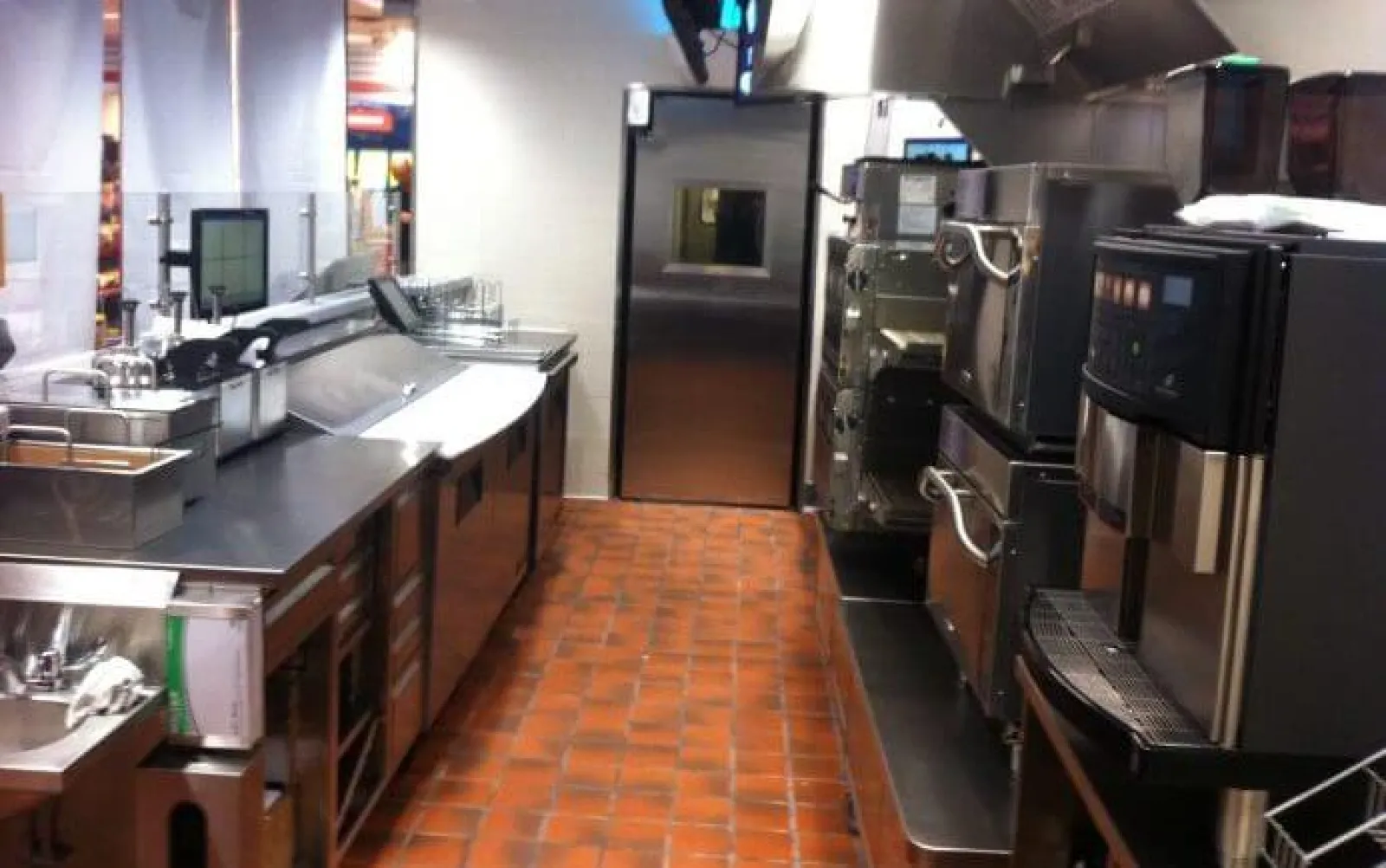 Commercial kitchen with stainless steel appliances, ovens, sinks, and terracotta tiled floor under bright lighting.