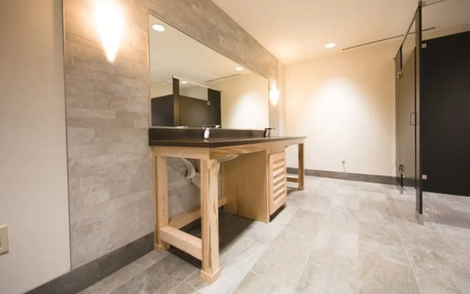 Modern public restroom with wooden vanity, large mirror, tiled floor, and black partition stalls under bright lighting