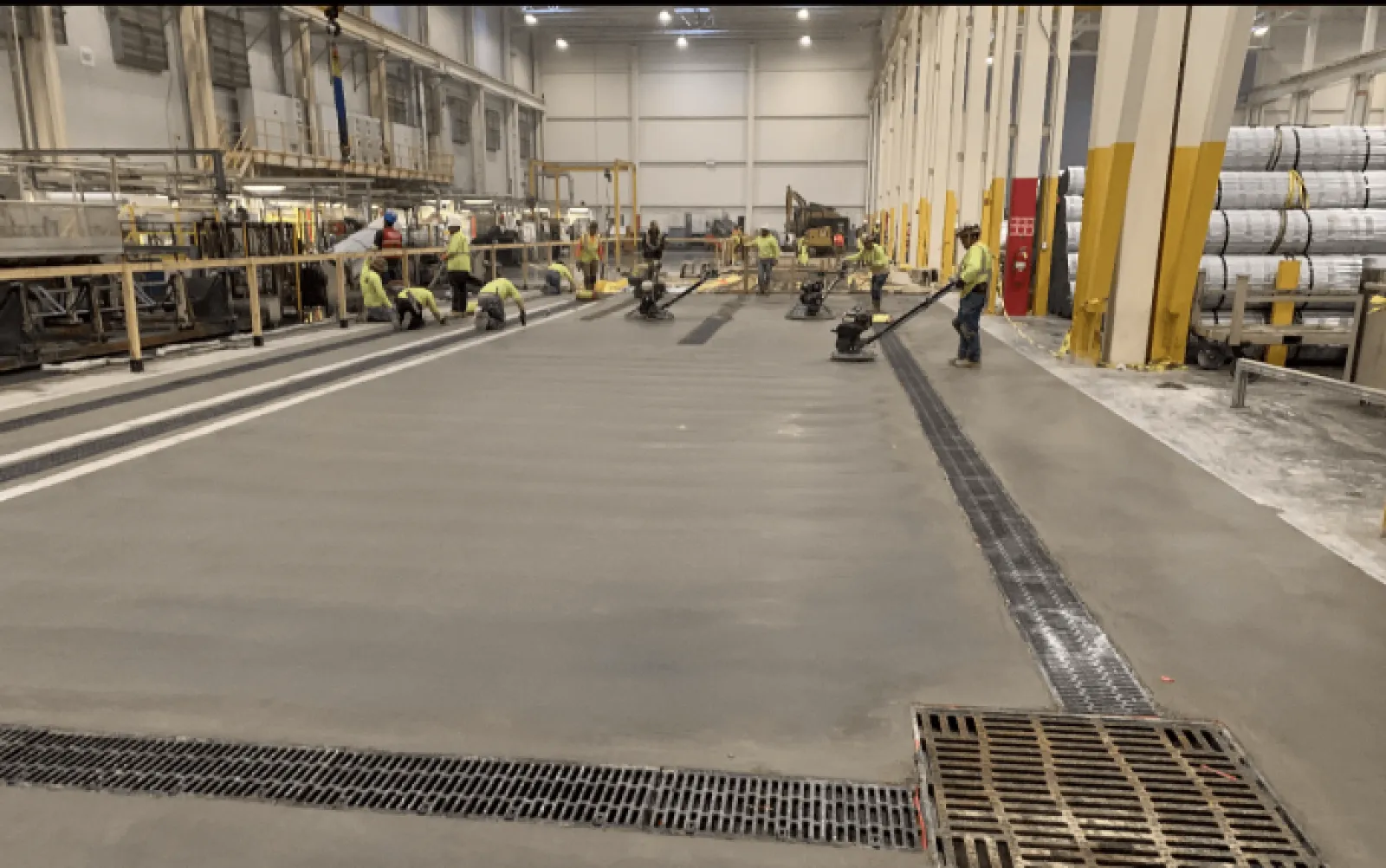 Workers smoothing and finishing a large concrete floor inside an industrial warehouse with heavy machinery.