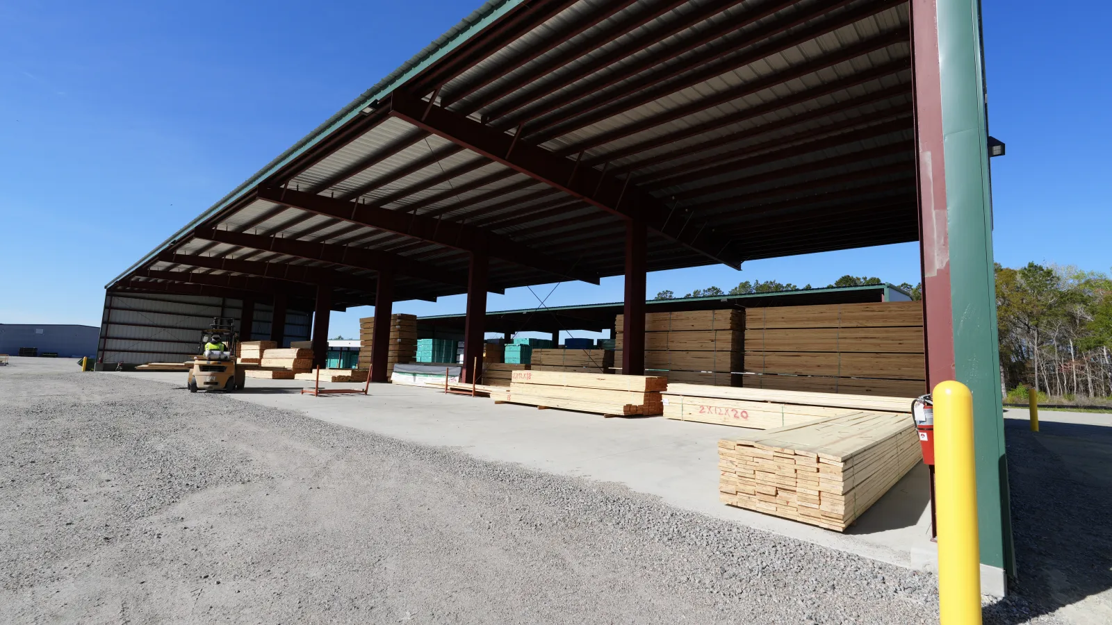 Outdoor lumber storage area with stacks of wooden boards under large metal shelter on sunny day.