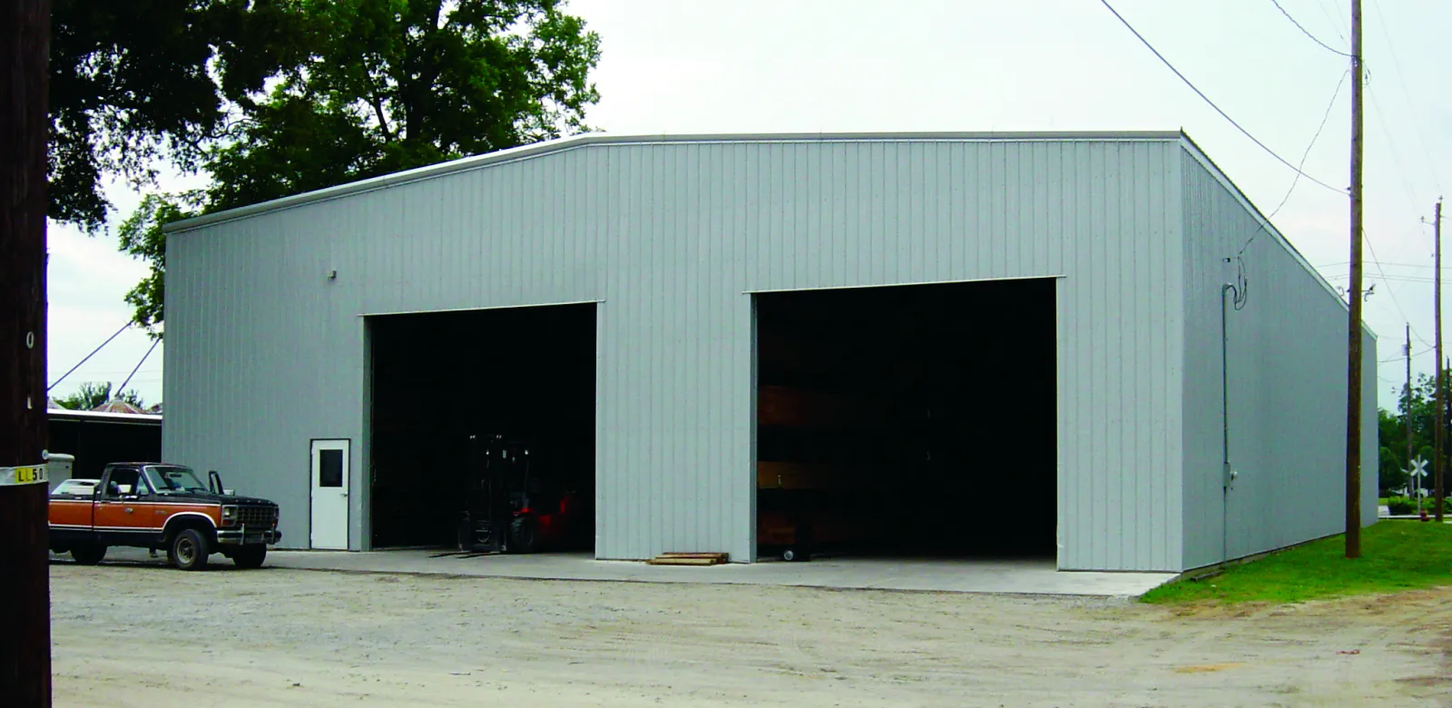 Large gray metal warehouse building with two open garage doors and a parked vintage truck outside under trees.