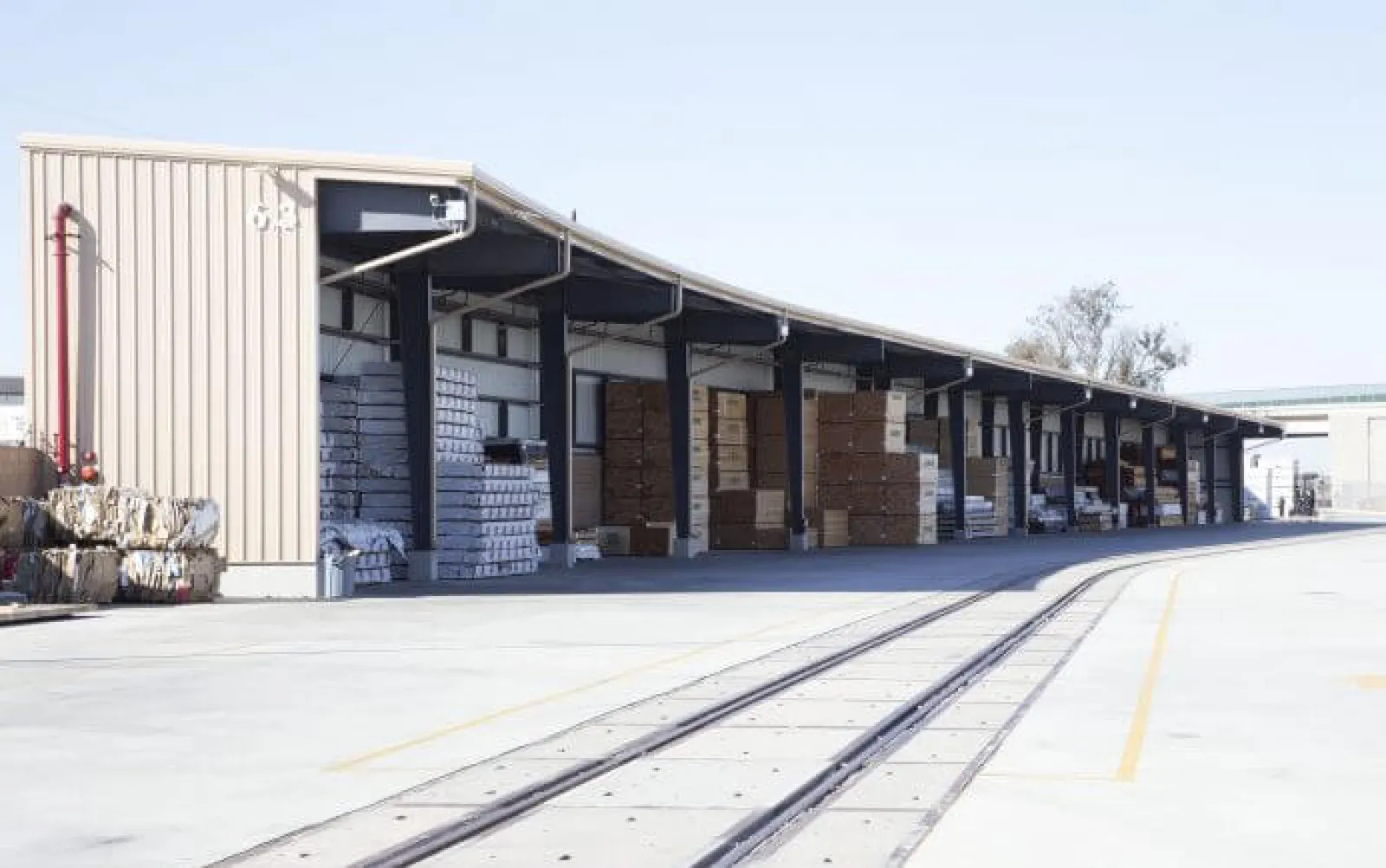 Outdoor warehouse with stacked boxes and pallets under a long covered loading dock near railway tracks.