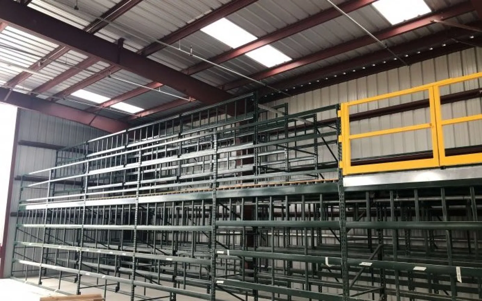 Empty metal storage racks inside a warehouse with a yellow safety gate on the upper platform under a skylit roof.
