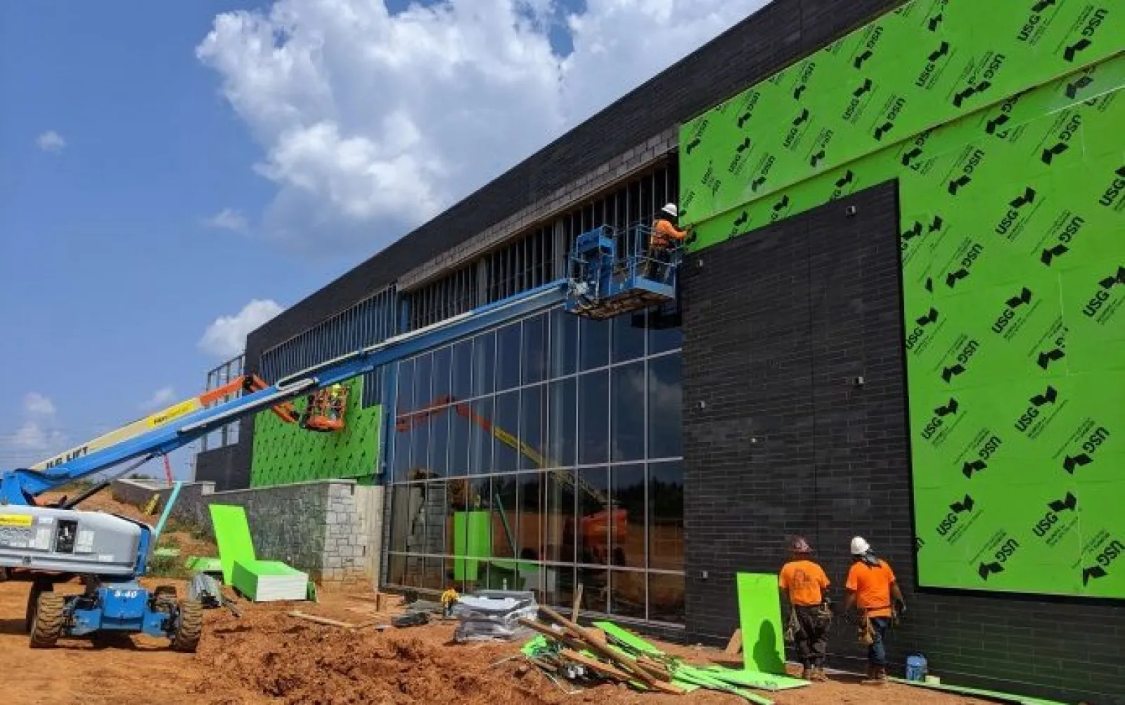 Construction workers install green sheathing and large windows on a commercial building under a partly cloudy sky.