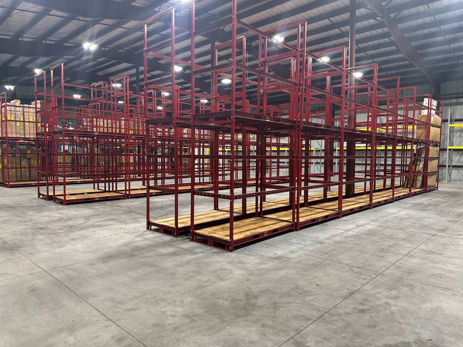 Empty red metal storage racks with wooden shelves inside a large warehouse with concrete floors and steel ceiling.