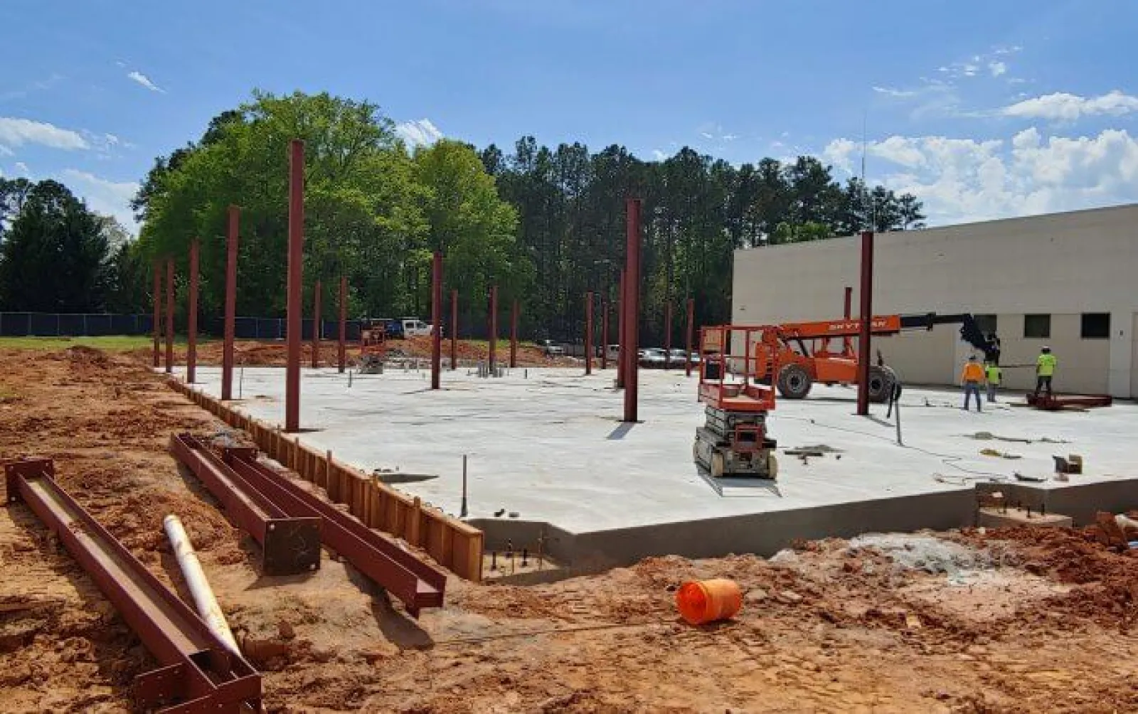 Construction site with steel beams and concrete foundation amid trees and workers under blue sky.