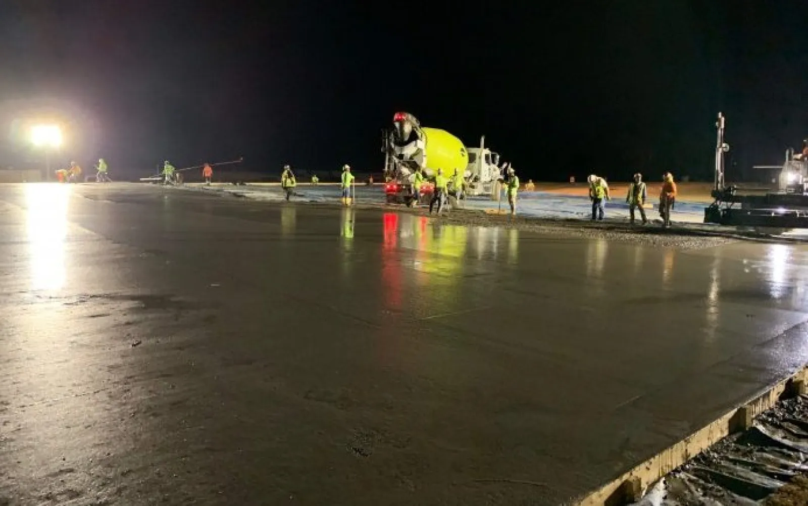 Nighttime construction crew pours and smooths concrete slab under bright lights with cement mixer truck present.