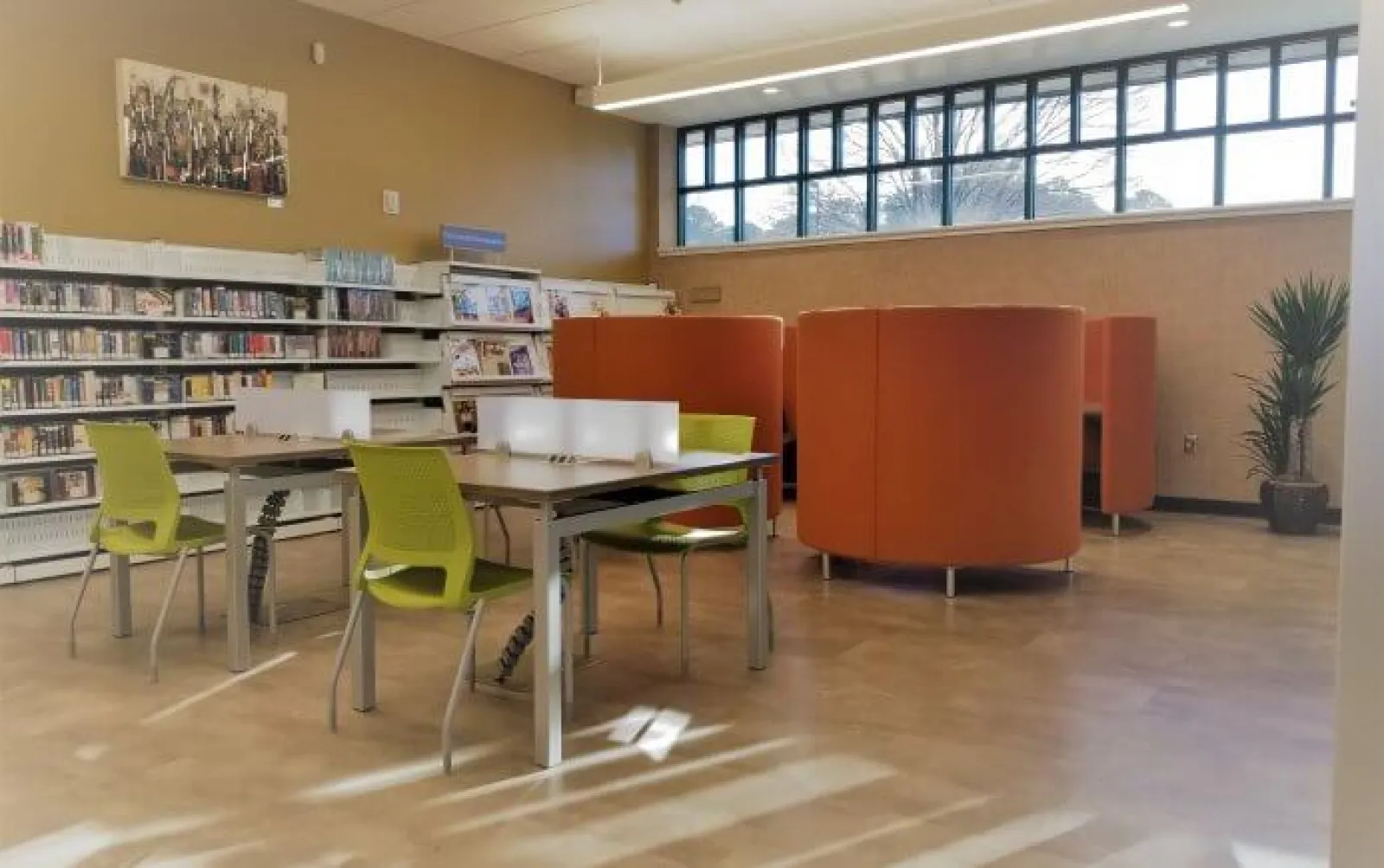 Modern library interior with green chairs, tables, orange curved booths, bookshelves, and large windows letting in natural light