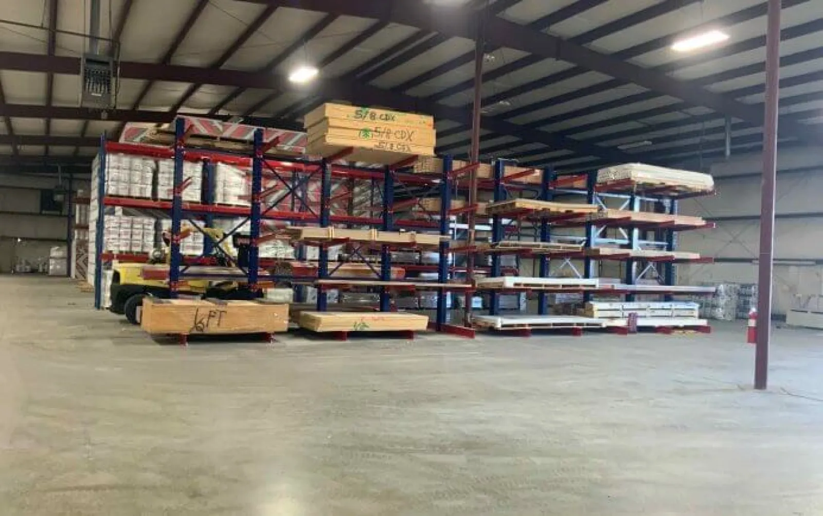 Warehouse interior with steel racks holding wooden planks and plywood sheets under fluorescent lights.