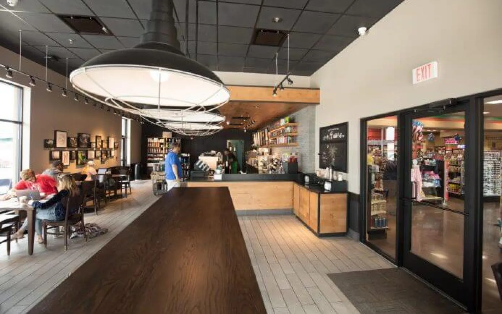 Modern coffee shop interior with wooden tables, large pendant lights, and customers sitting near windows.