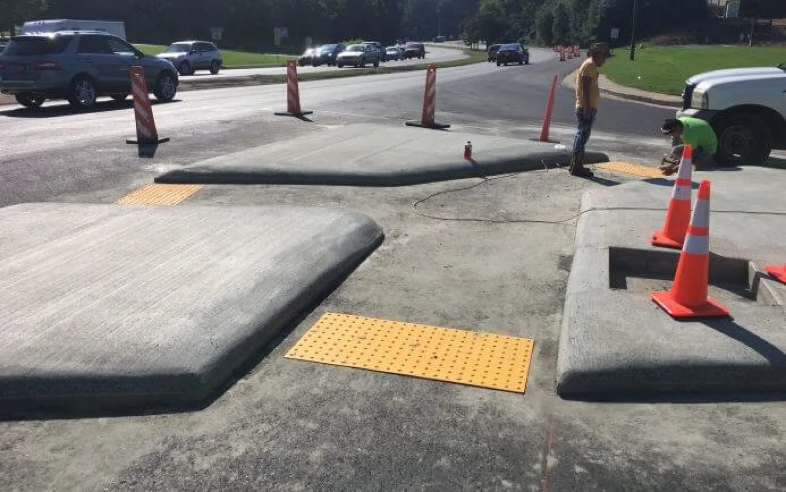 Newly constructed concrete curb ramps with tactile paving and traffic cones near a busy road under clear sky.