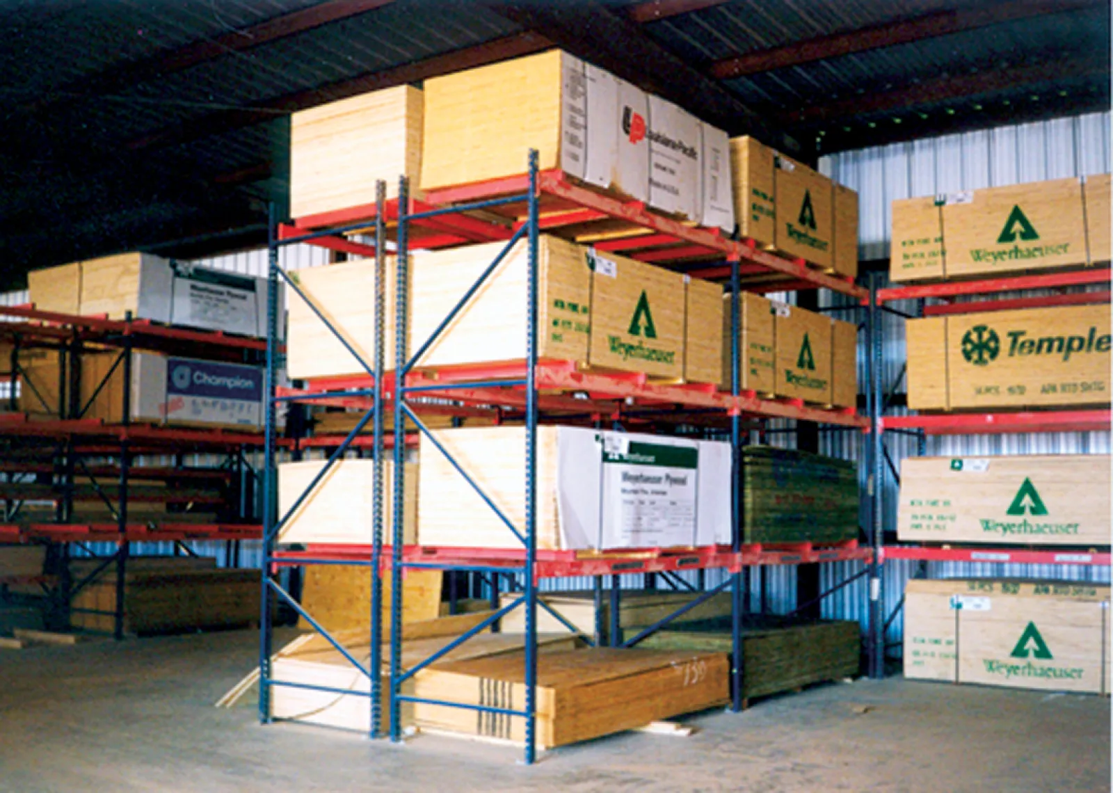 Warehouse interior showing stacks of lumber on metal racks under a corrugated roof with branded packaging