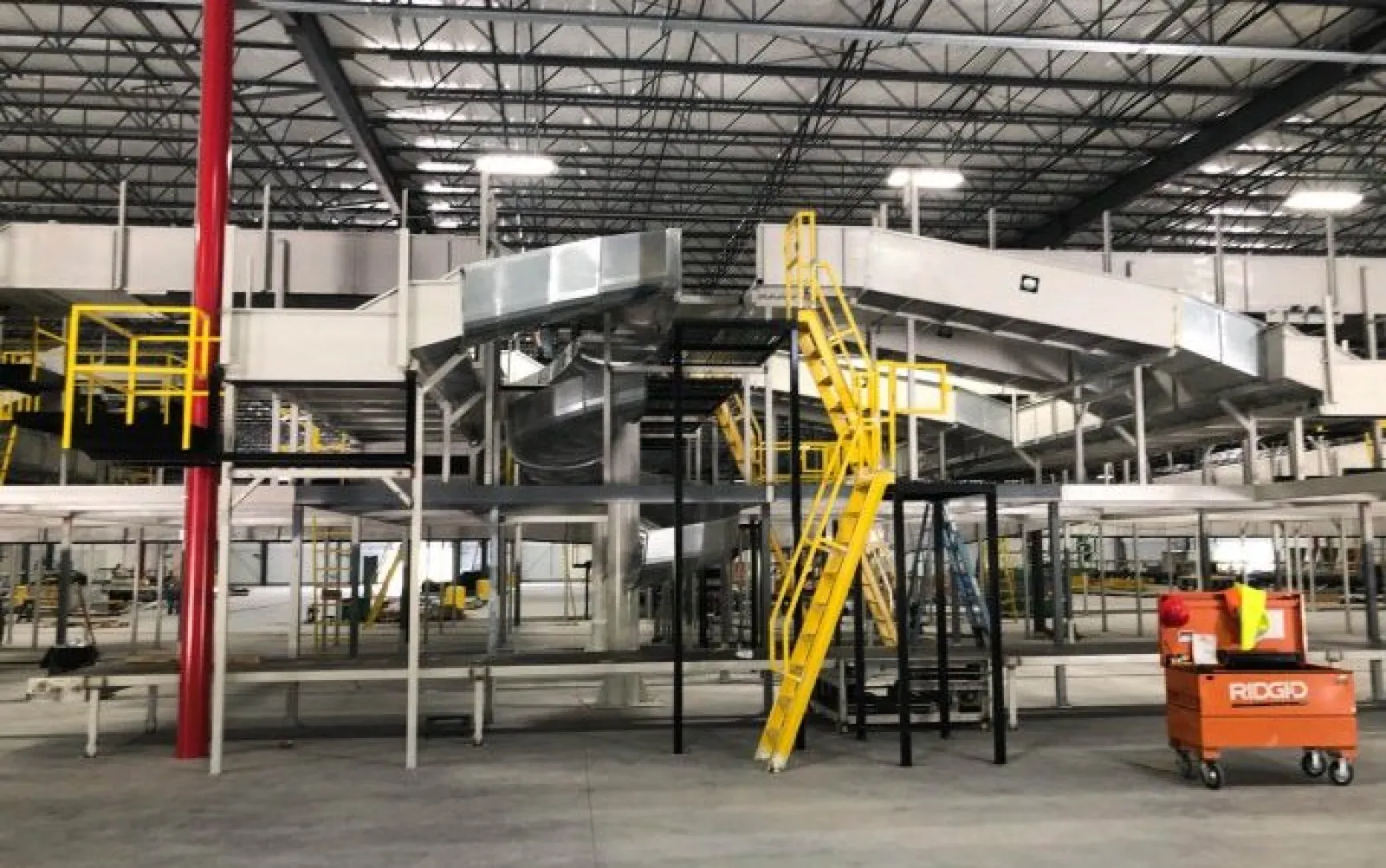 Industrial warehouse with conveyor belts, yellow stairs, and RIDGID tool cart on concrete floor under metal roof