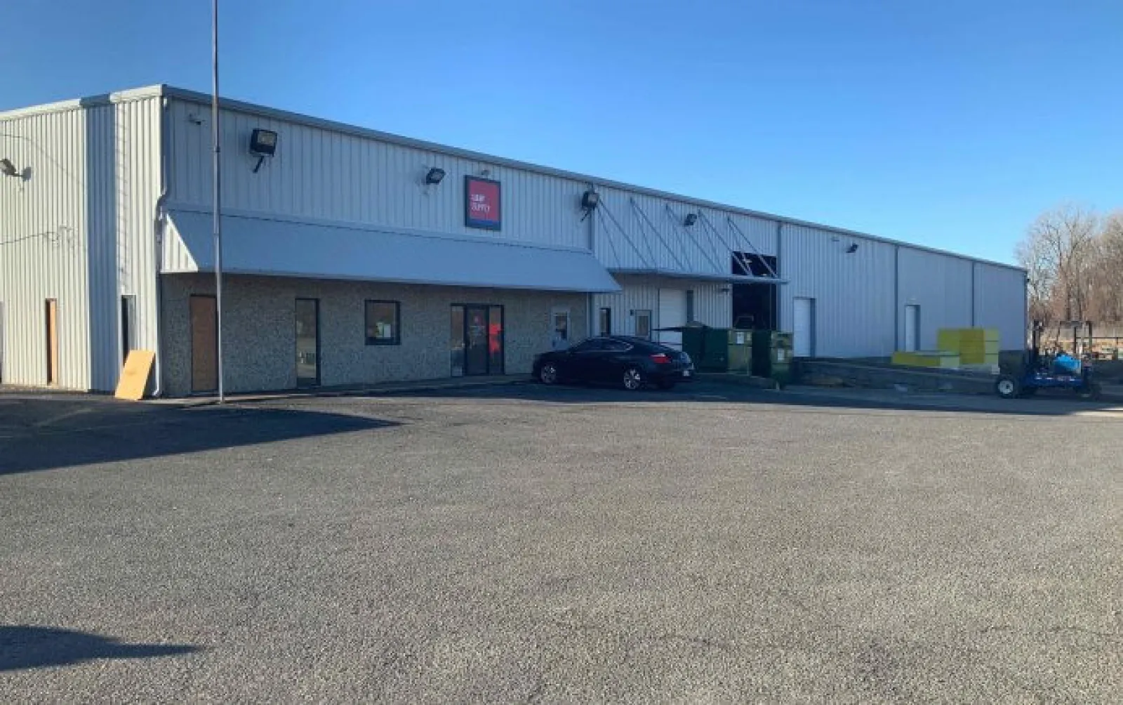 Large industrial warehouse with metal siding under clear blue sky and a parked black car outside.