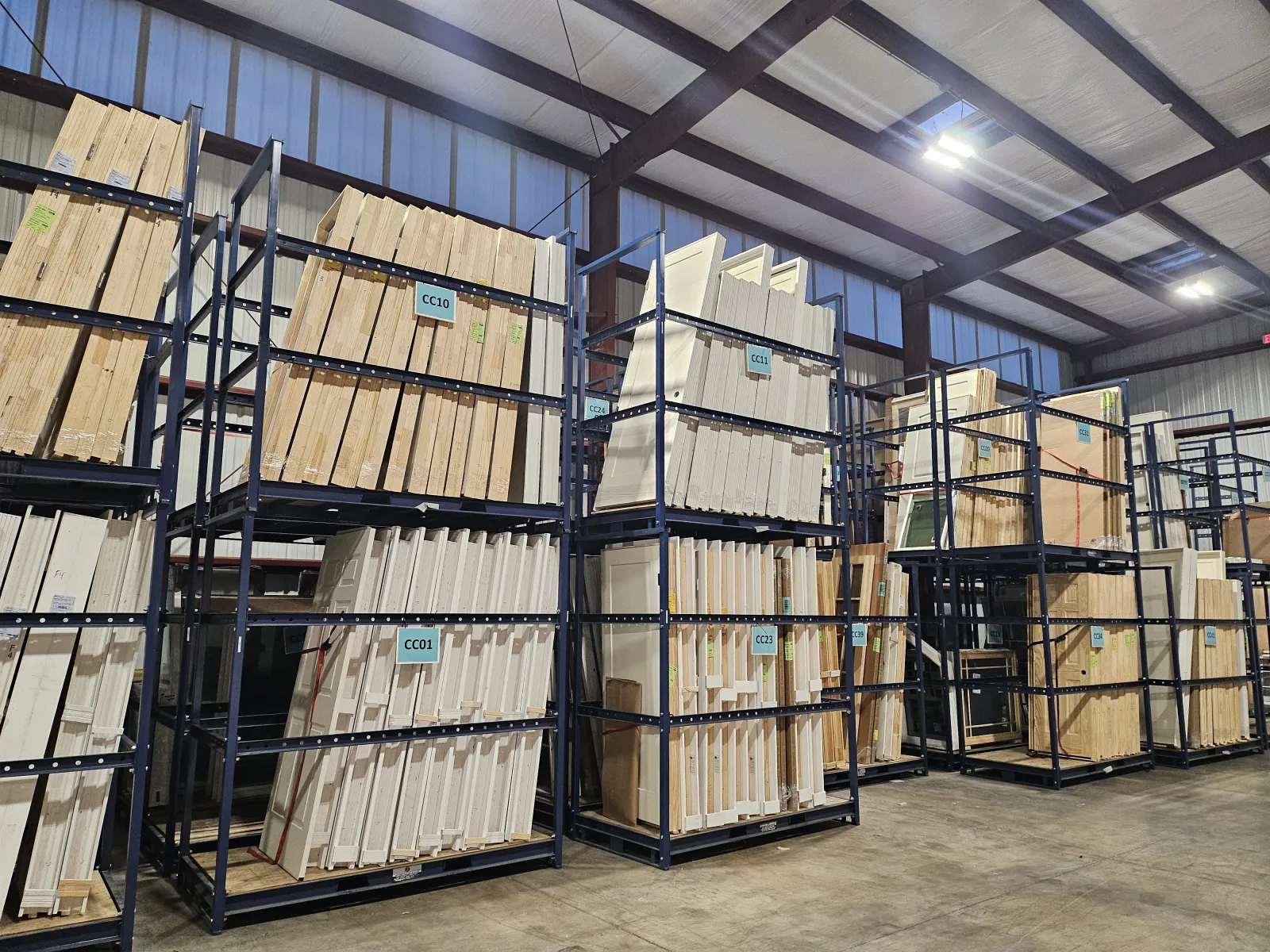 Wood panels and lumber stacked on tall metal racks in a spacious warehouse with concrete floors and high ceilings.