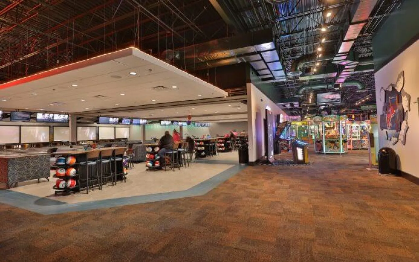 Spacious modern bowling alley with seating area, bowling balls, and arcade games under industrial ceiling.