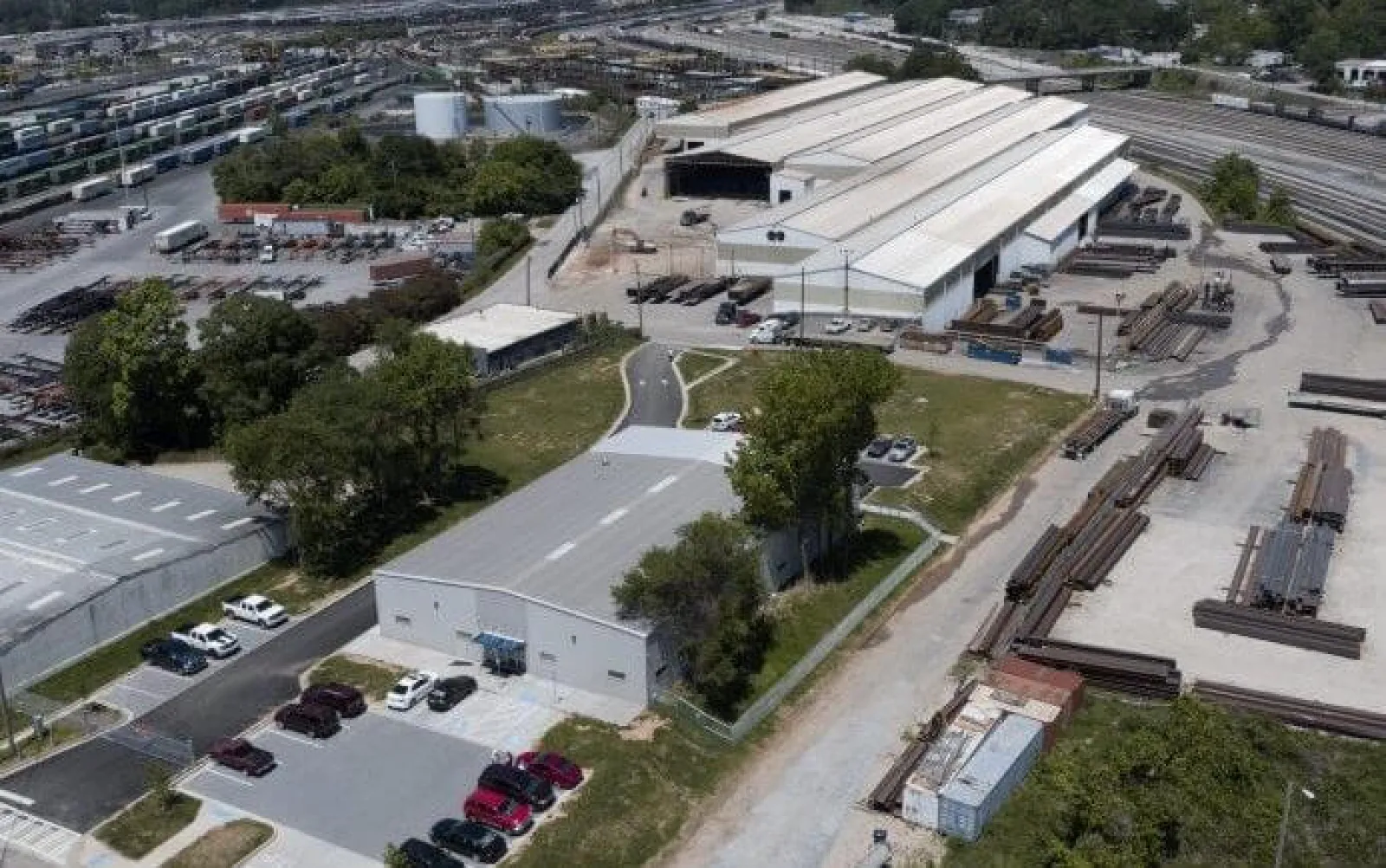 Aerial view of an industrial facility with warehouses, parked cars, stacks of metal beams, and railway tracks.
