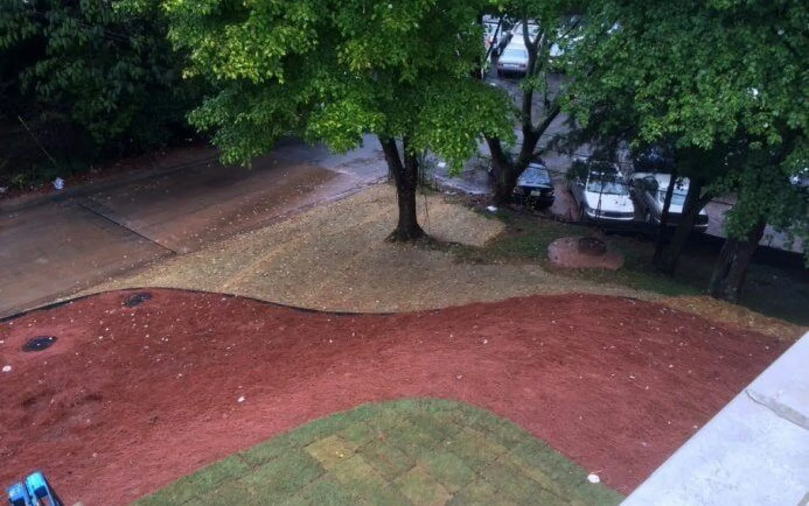 Curved red dirt pathway surrounded by grass and trees with parked cars in the background in an outdoor area.