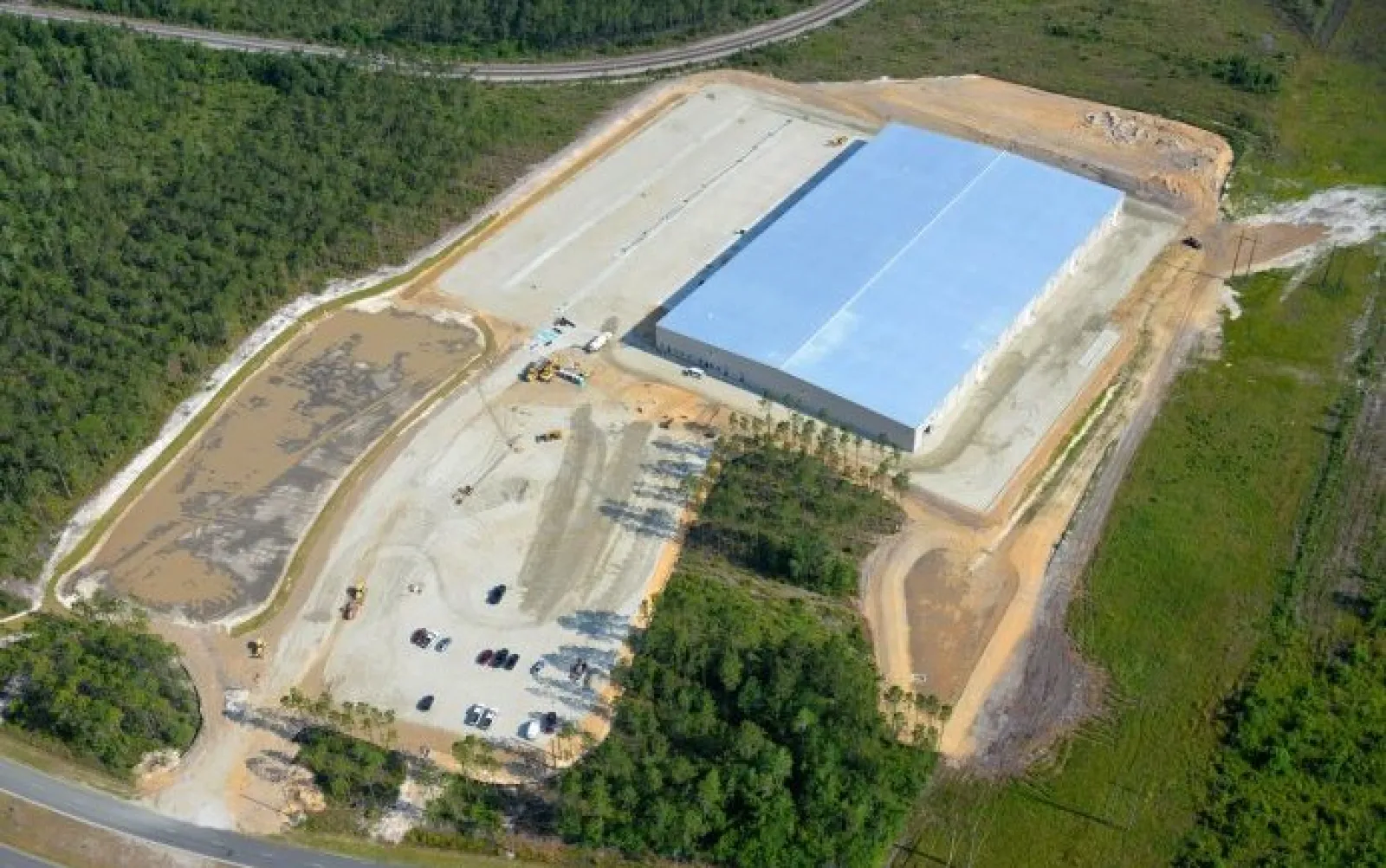 Aerial view of a large warehouse under construction surrounded by cleared land and forested areas.