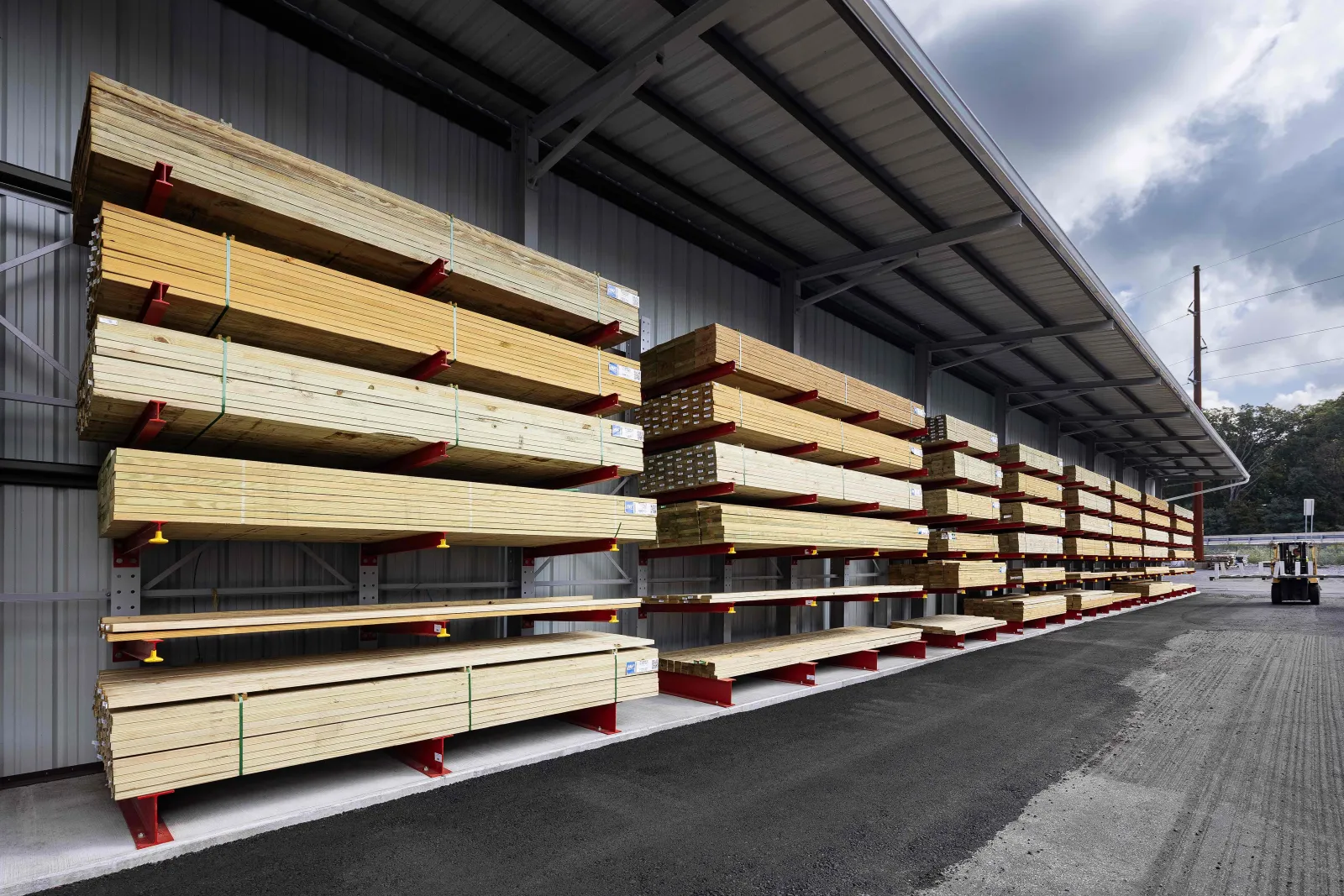 Outdoor lumber storage racks filled with stacked wooden planks under a metal canopy on a paved surface.
