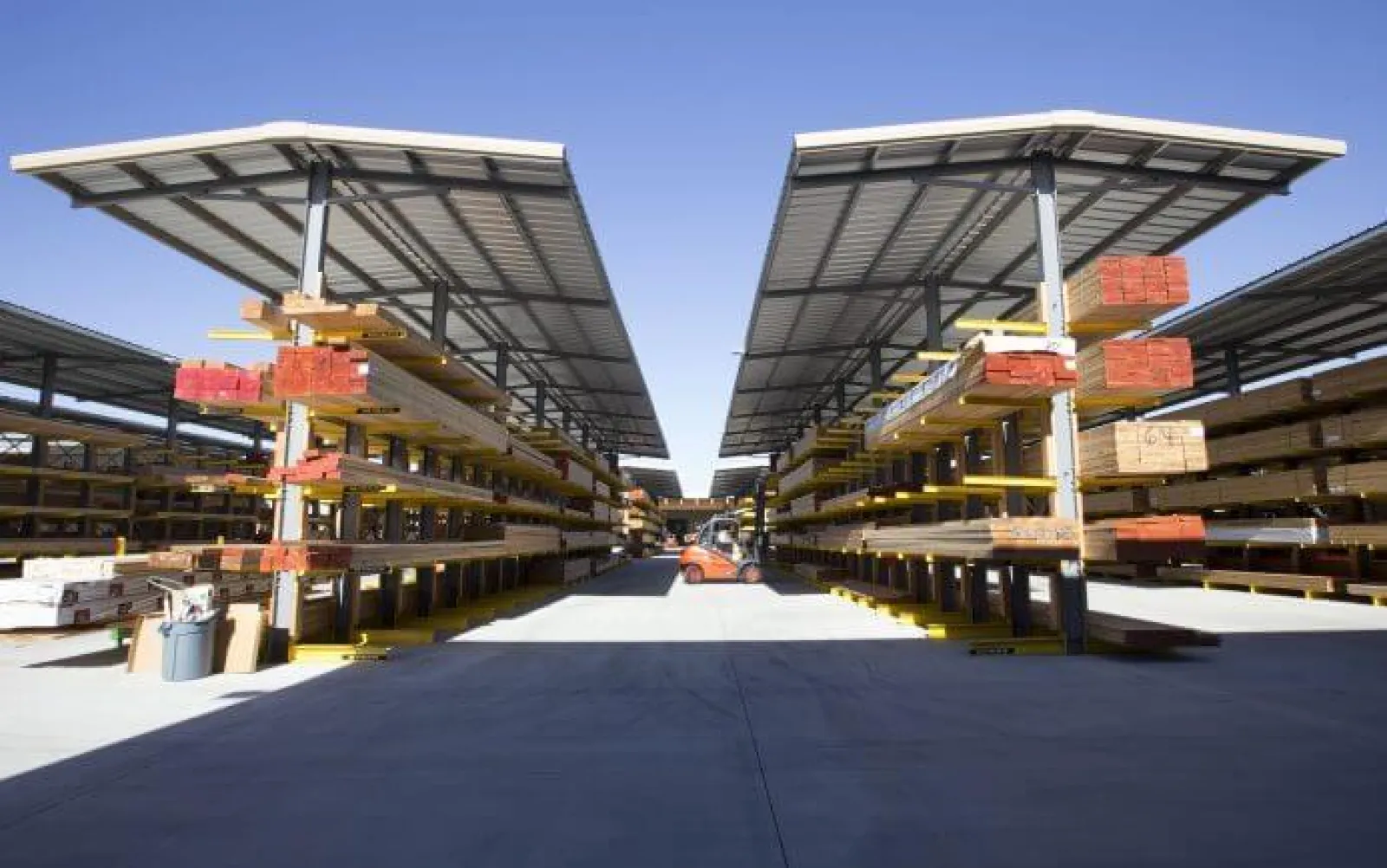 Outdoor lumber yard with organized stacks of wood under metal canopies and a forklift in the center aisle.