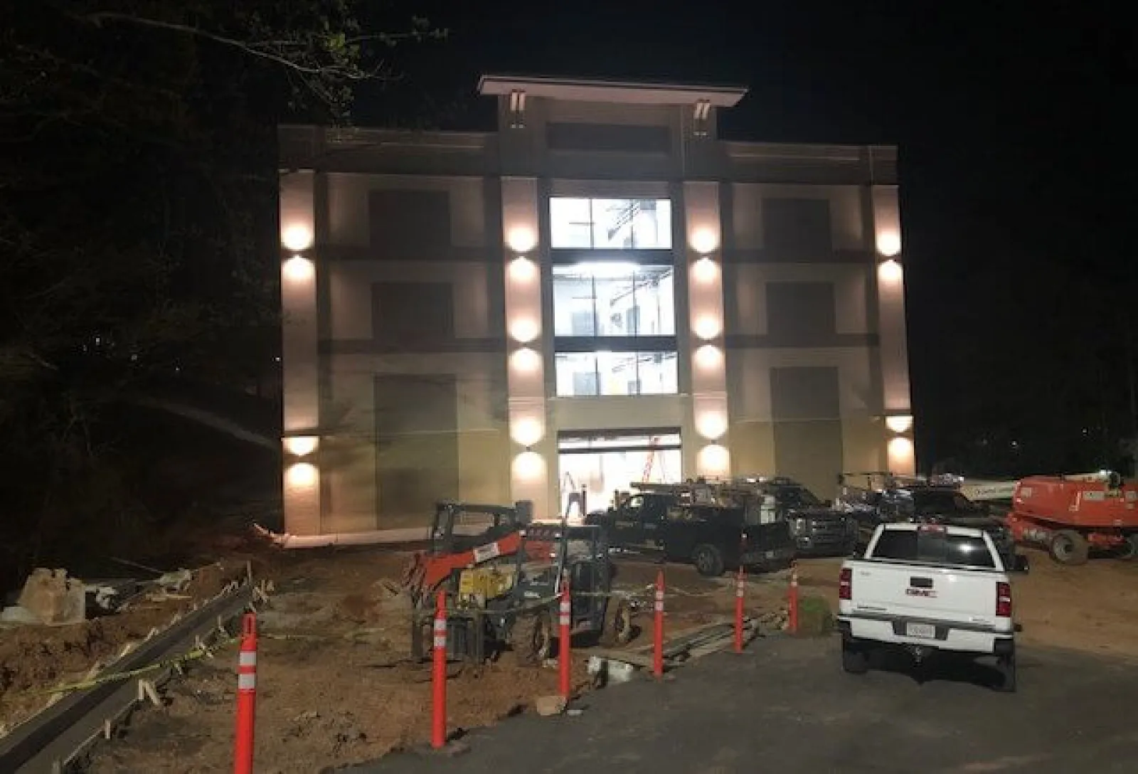 Illuminated building under construction at night with parked trucks and construction equipment.