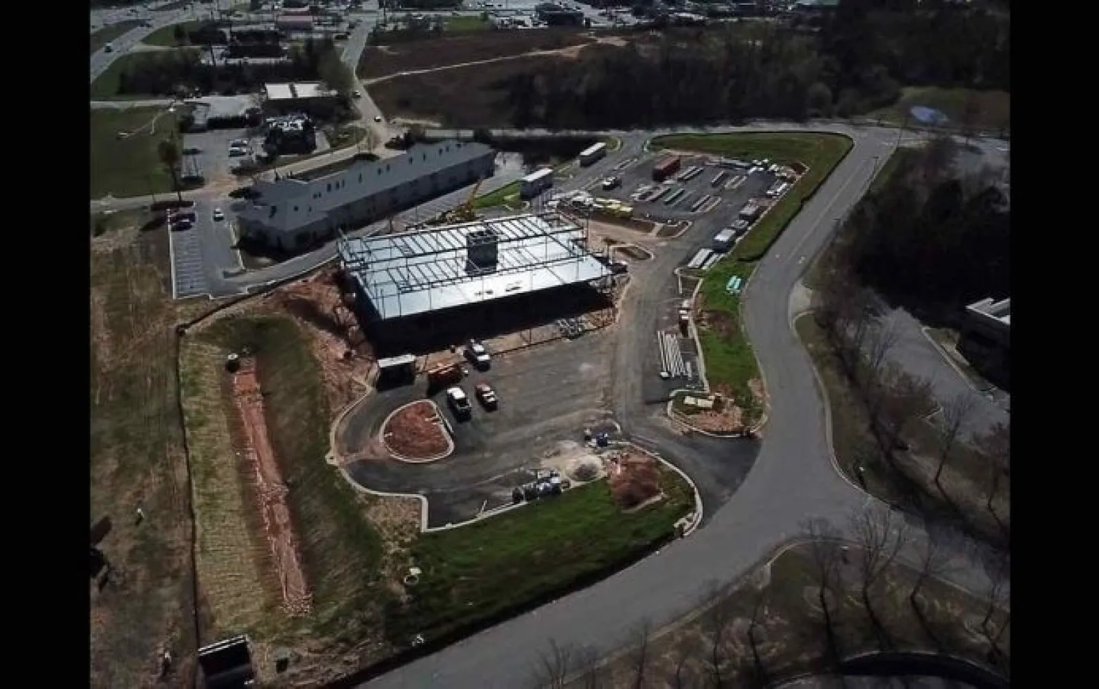 Aerial view of a construction site with a building framework, surrounding roads, parking, and vehicles during daytime.