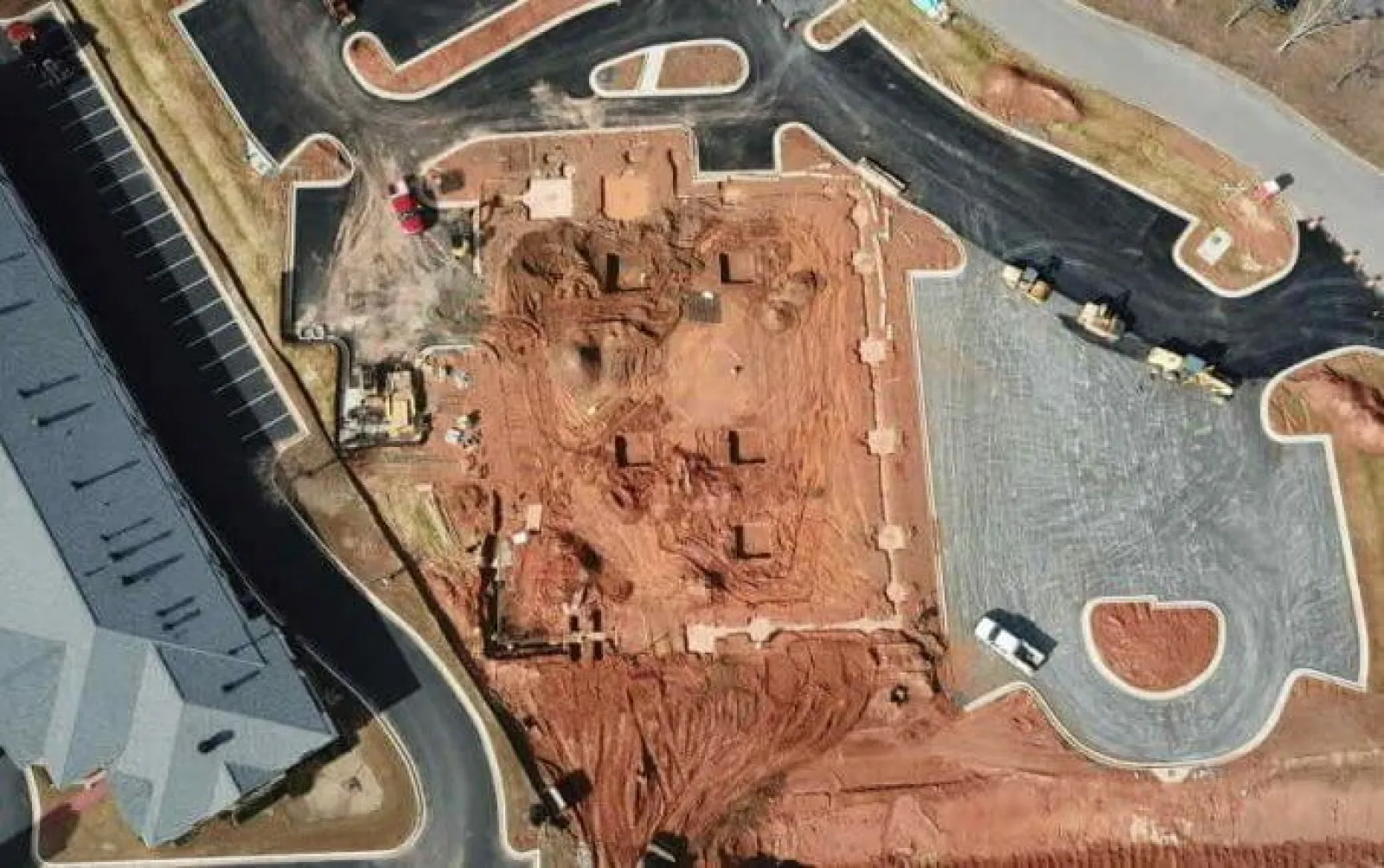 Aerial view of a construction site with cleared land, construction vehicles, paved parking, and surrounding roads.