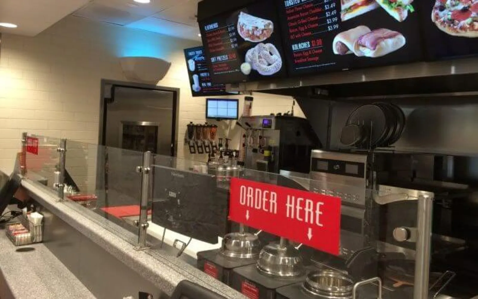 Counter and menu screens inside a modern sandwich shop with an order here sign and clear protective barriers.