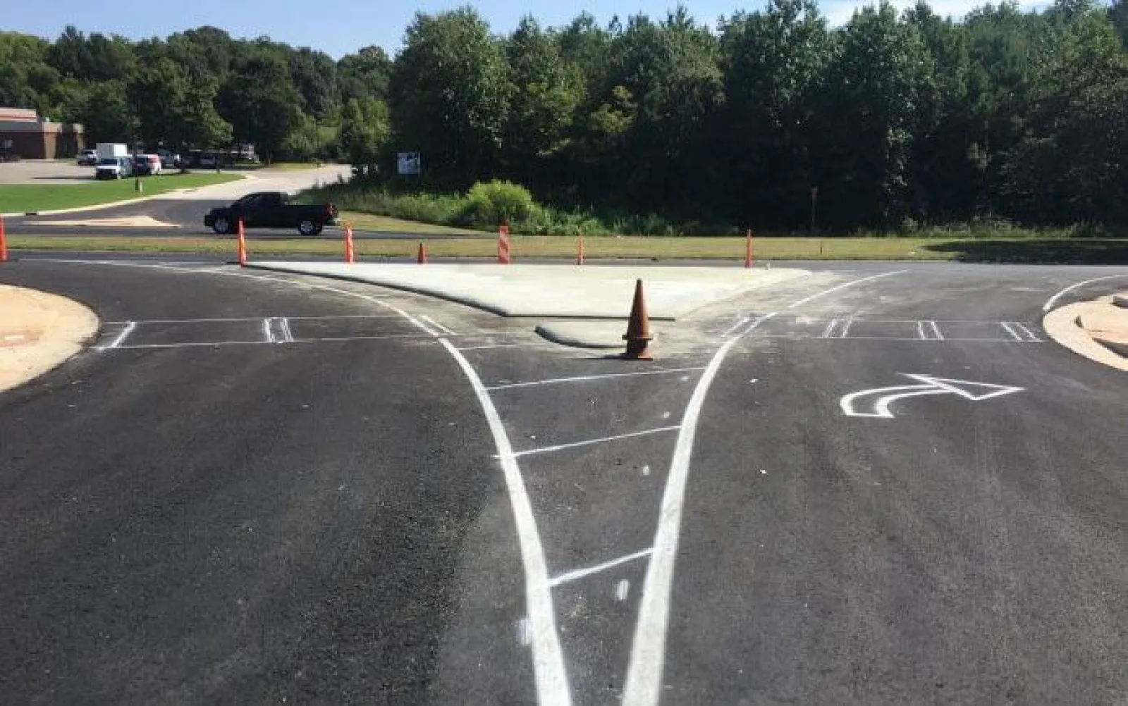 Freshly paved road with new white lane markings and a traffic cone at a fork under clear skies with trees.