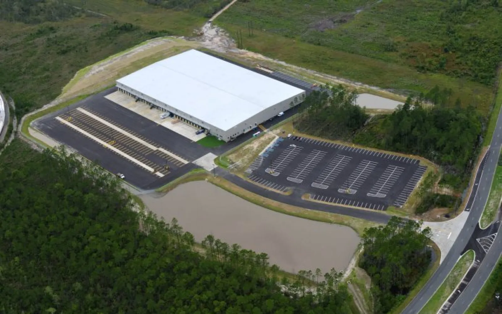 Aerial view of a large white-roof warehouse surrounded by parking lots, a pond, and green wooded areas.