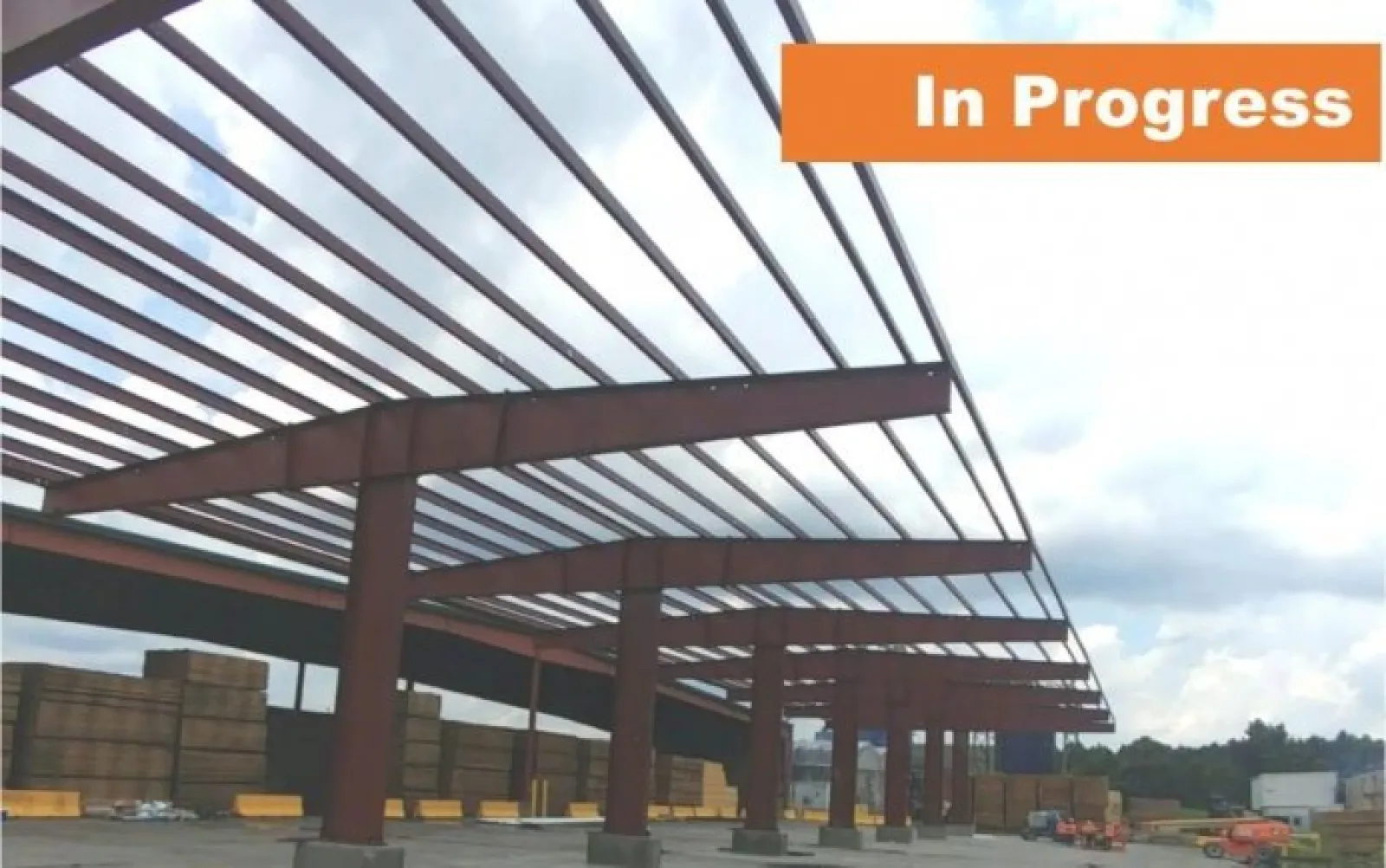 Steel frame construction of a large building in progress under a cloudy sky with stacked wood and equipment nearby